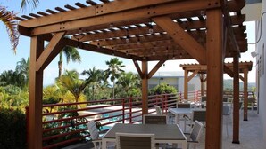 Une pergola en bois, agrémentée de tables et de chaises, qui offre une vue sur une piscine et des palmiers.
