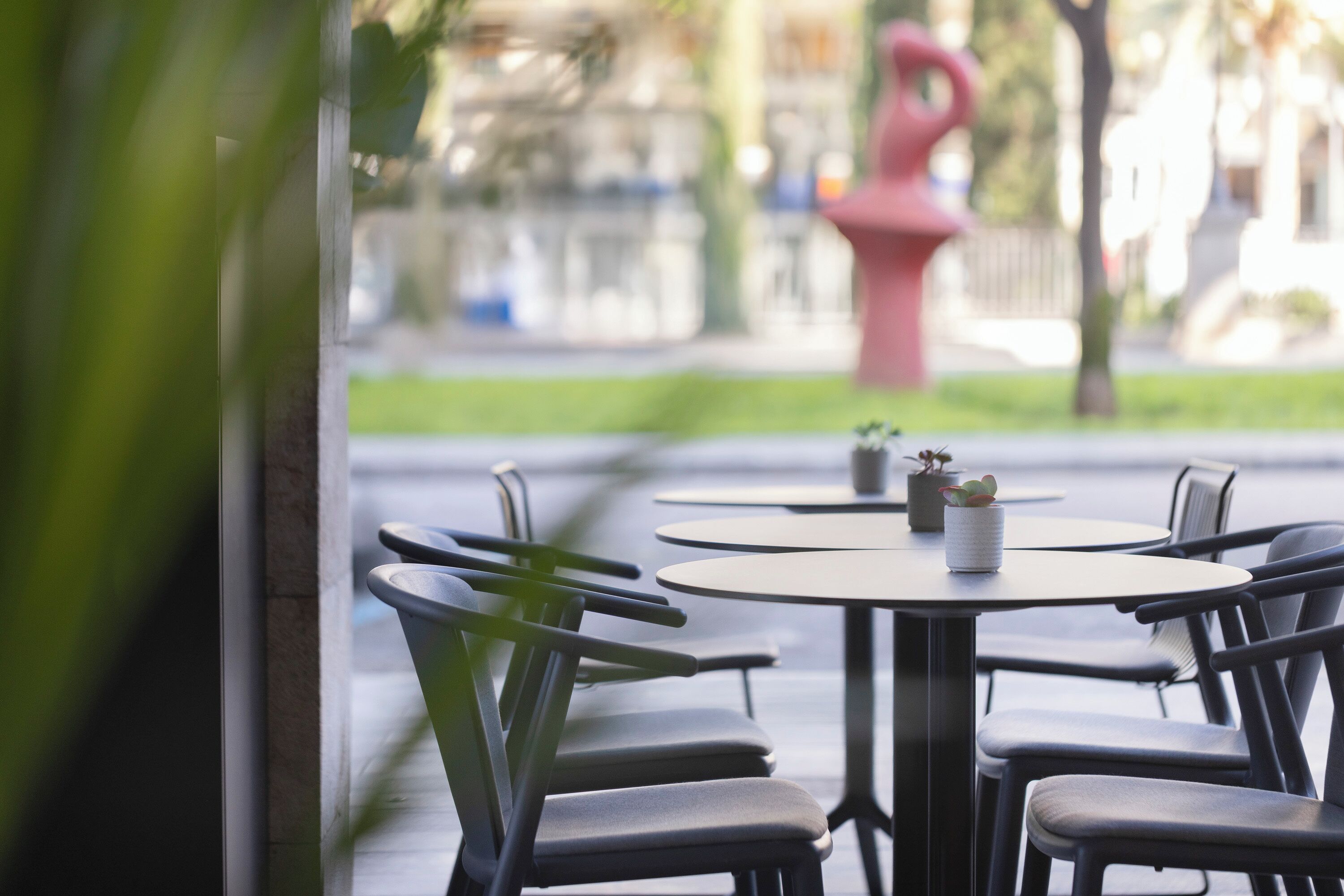 A row of round tables with chairs, a potted plant, and a red sculpture in the background.