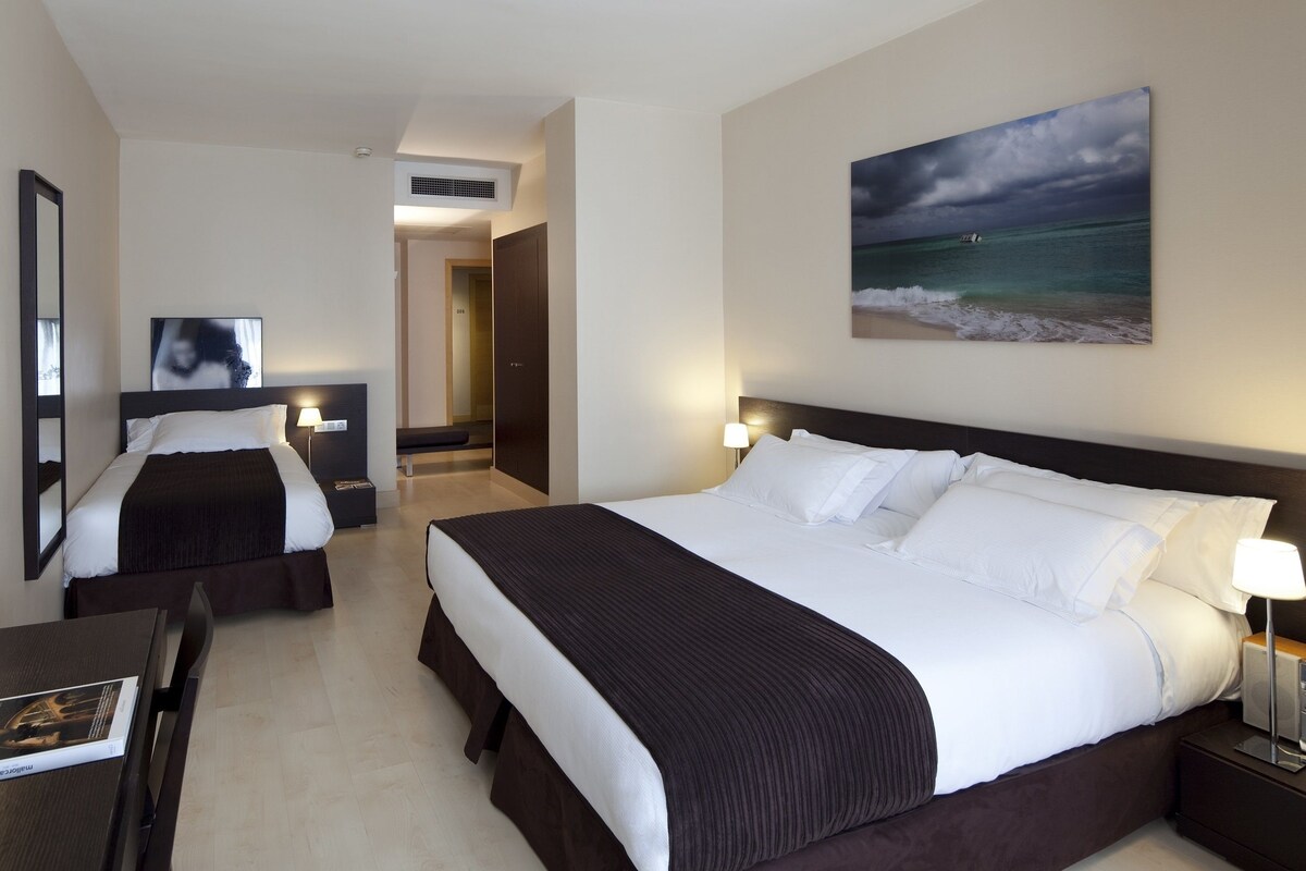 A hotel room with two beds, a desk, a chair, a painting, and a view of the sea.