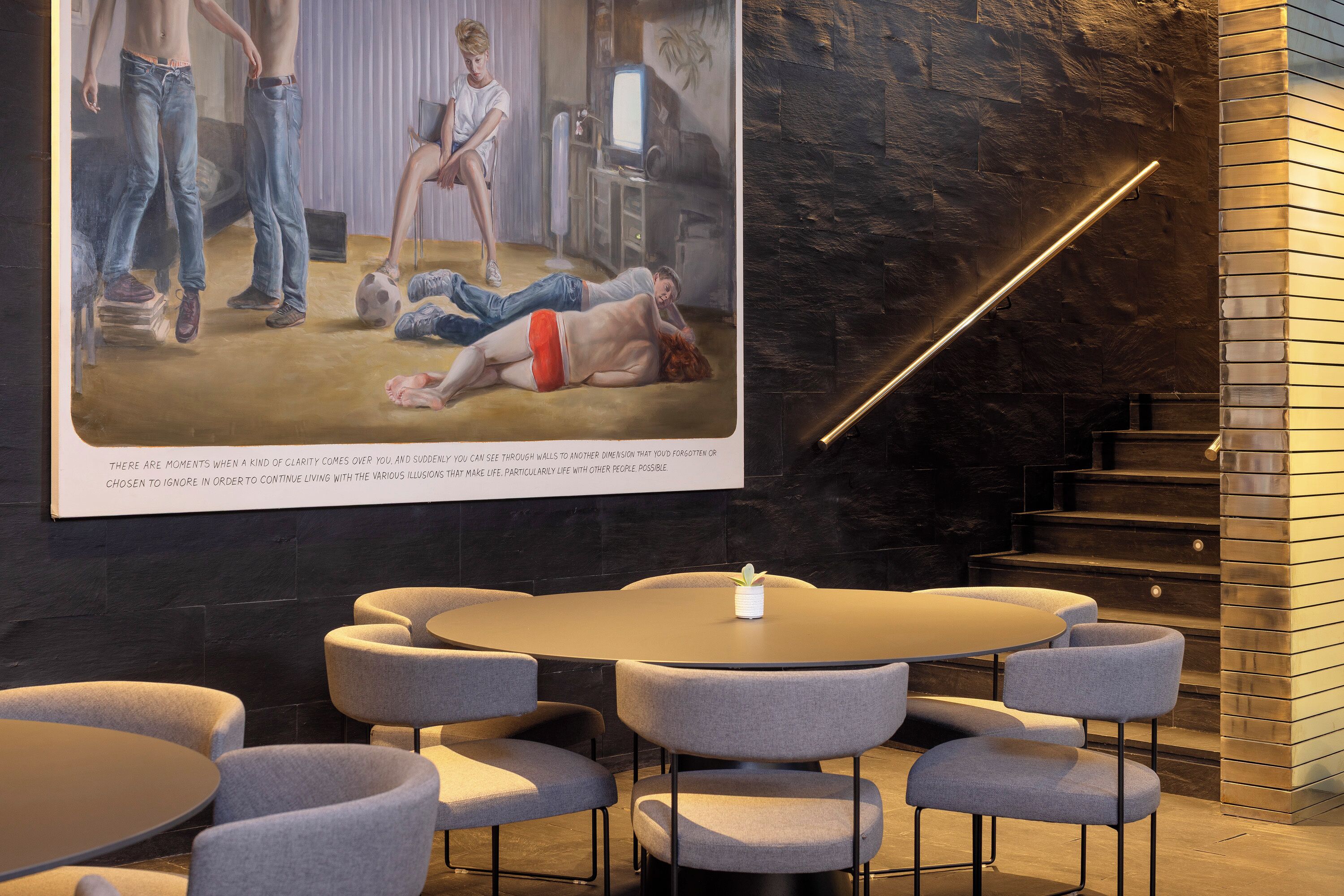 Un interior moderno con una mesa redonda y sillas, una escalera y una obra de arte grande enmarcada que muestra una escena con gente y un perro.