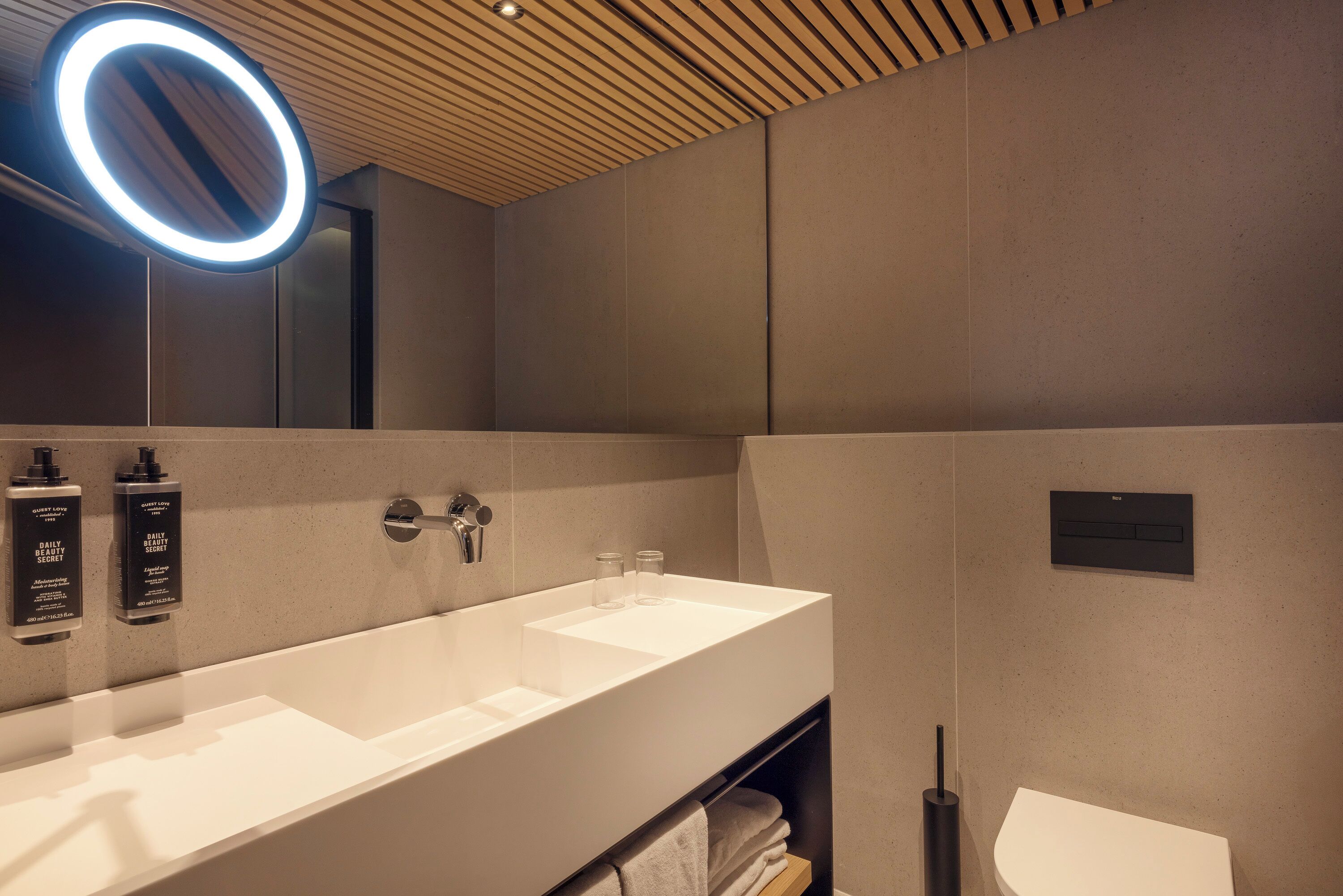 A modern bathroom with a large white sink, a mirror with a circular light, and a towel rack.