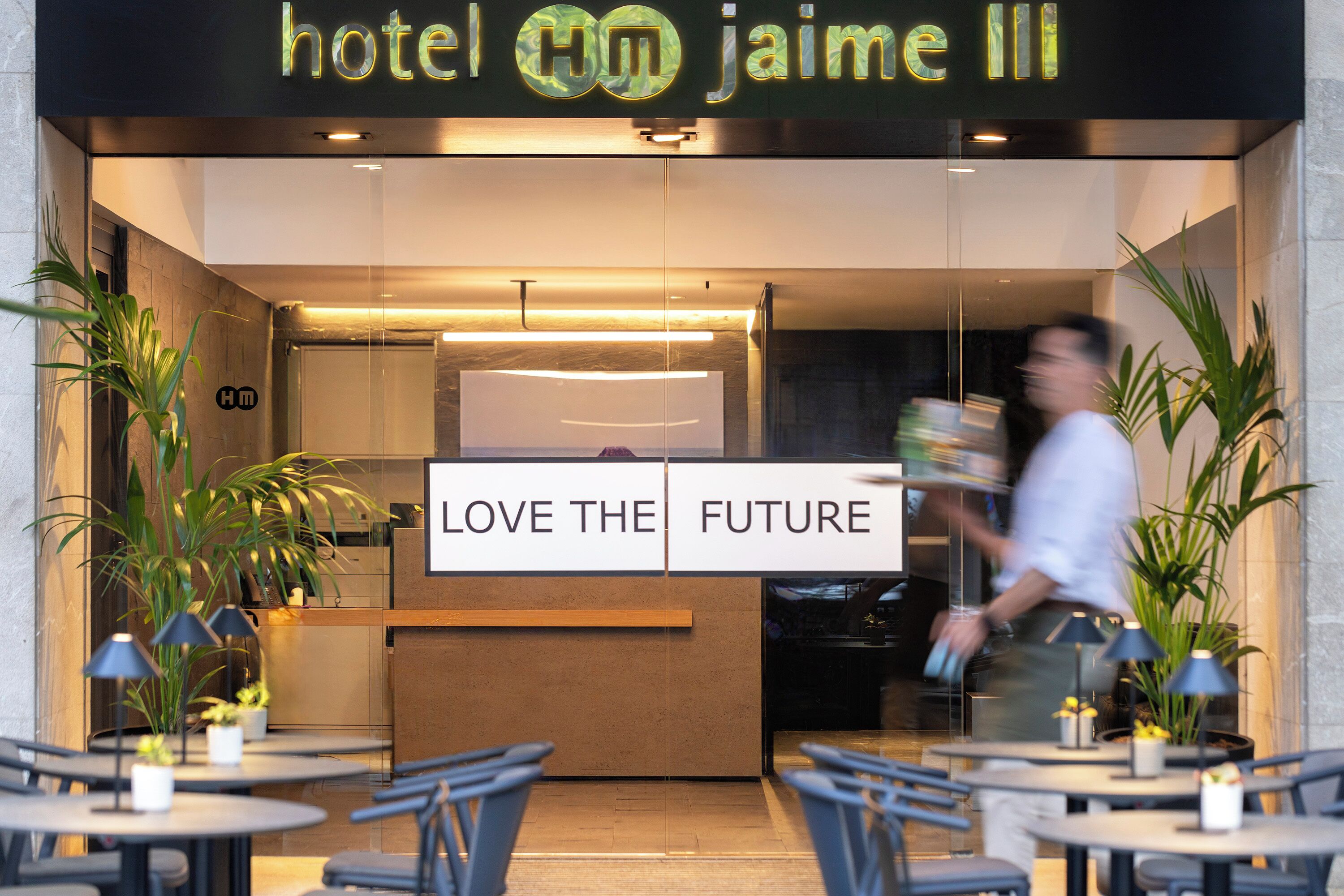 Entrada de hotel con mostrador de recepción, un letrero que dice "LOVE THE FUTURE" y zona exterior con mesas y sillas.