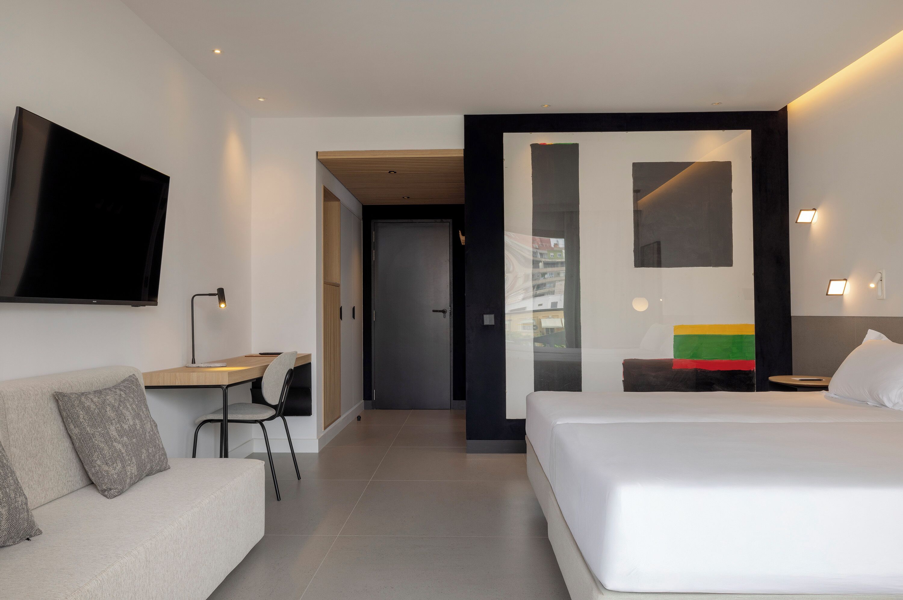 Una habitación de hotel moderna con cama, escritorio y sofá. Hay un televisor montado en la pared y una puerta que da a otra habitación.