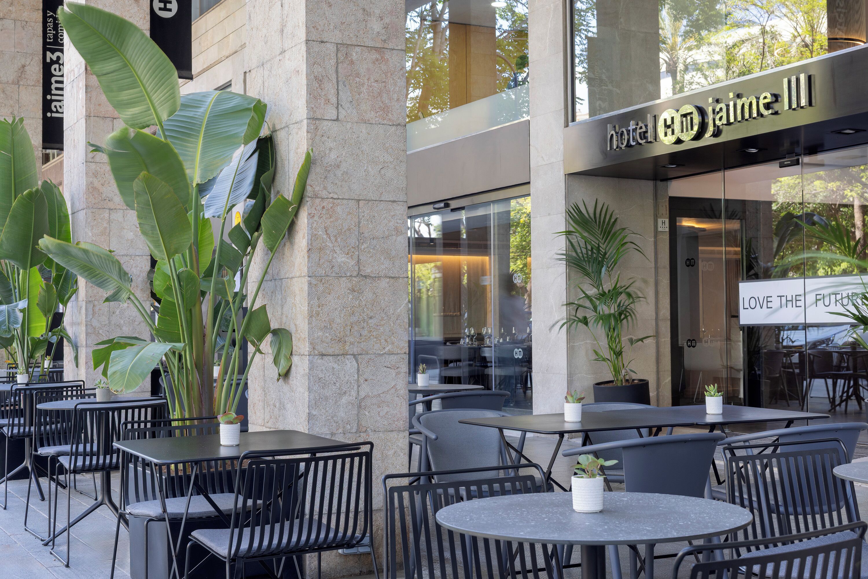 Un espace extérieur aménagé avec des tables et des chaises, des plantes en pot et l’entrée de l’hôtel, reconnaissable à son panneau.