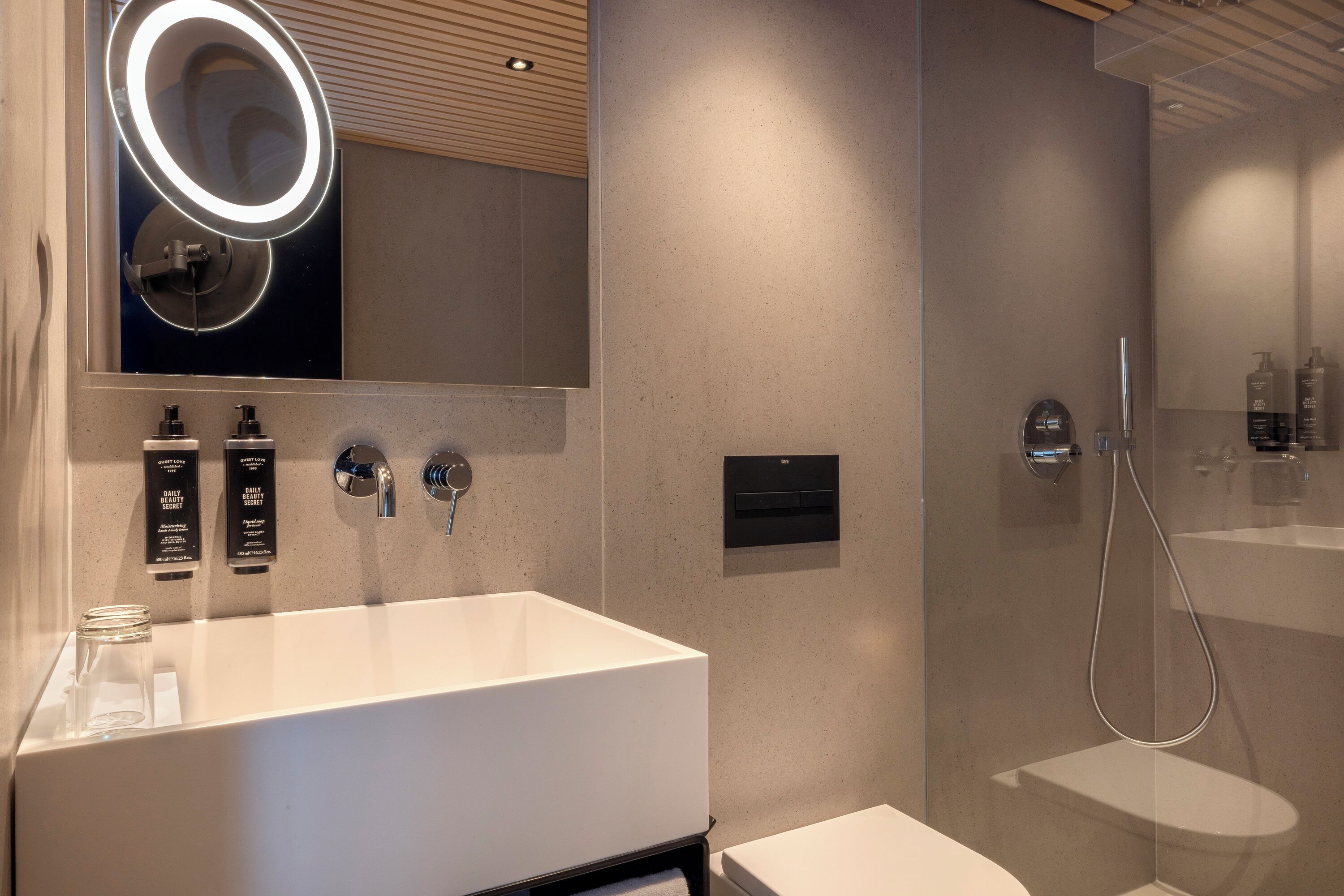 Un baño moderno con un lavamanos grande de color blanco, un espejo con luz circular y una zona de ducha con mampara de vidrio.