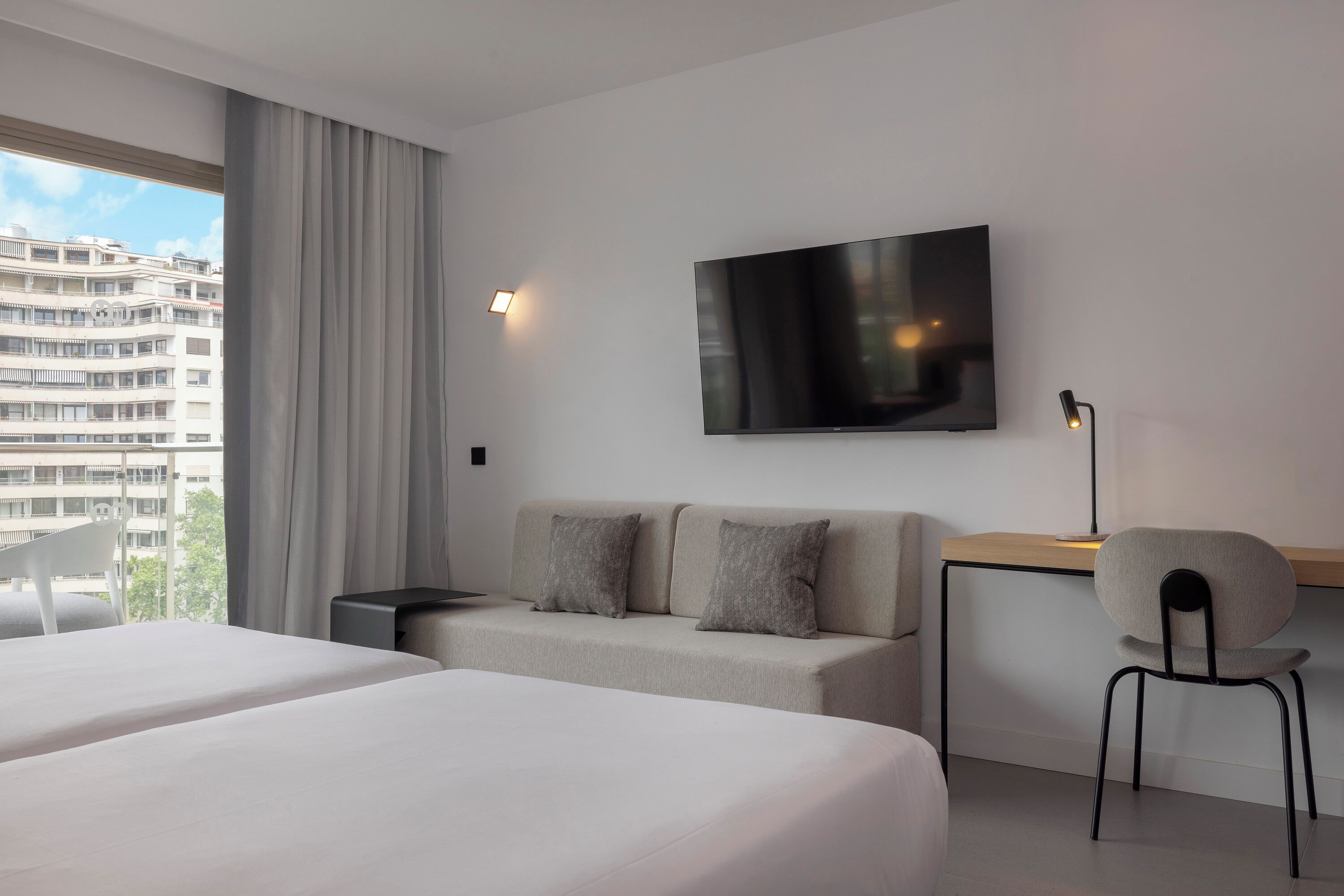 Habitación de hotel moderna con una cama grande, un sofá, un escritorio y una silla. Hay un televisor en la pared y una ventana con cortinas.