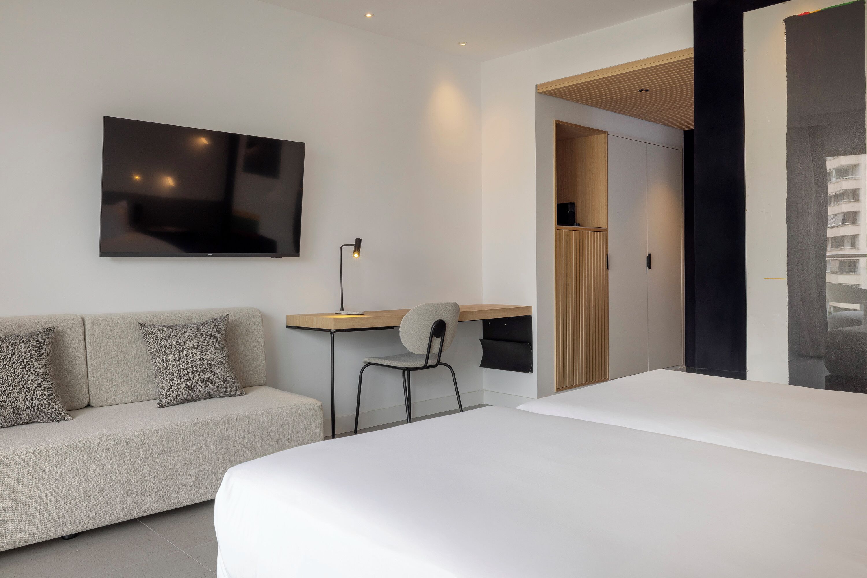 A modern hotel room with a large bed, a sofa, a desk with a chair, a flat-screen TV mounted on the wall, and a view of the city through a large window.
