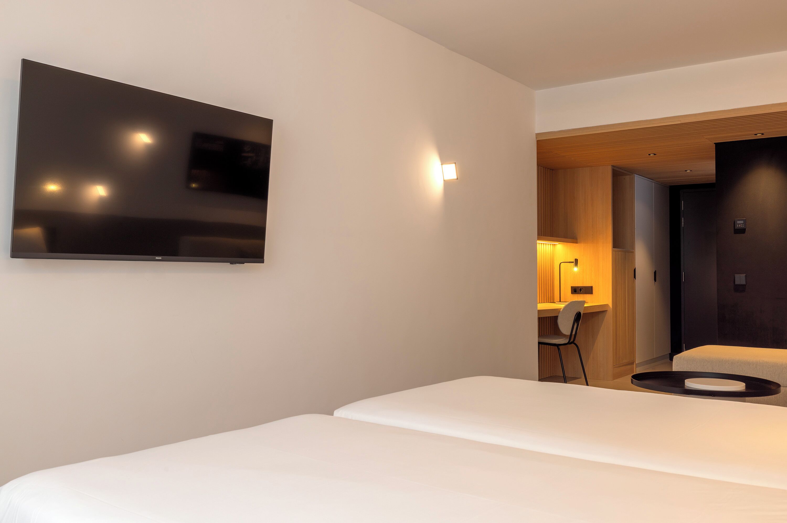 A modern hotel room with a flat-screen TV mounted on the wall, a bed with a circular bedside table, and a desk area with a chair.