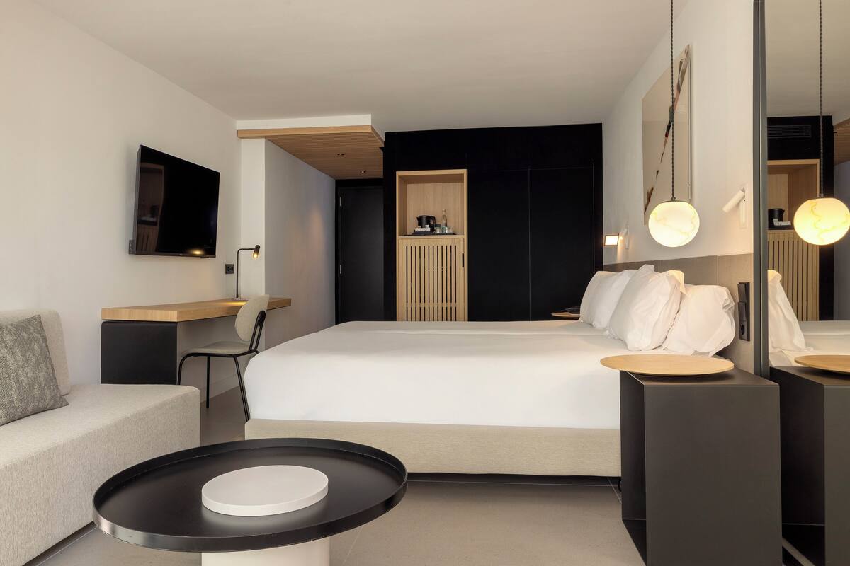 A modern hotel room with a large bed, a desk, a chair, a small table, and a TV mounted on the wall.