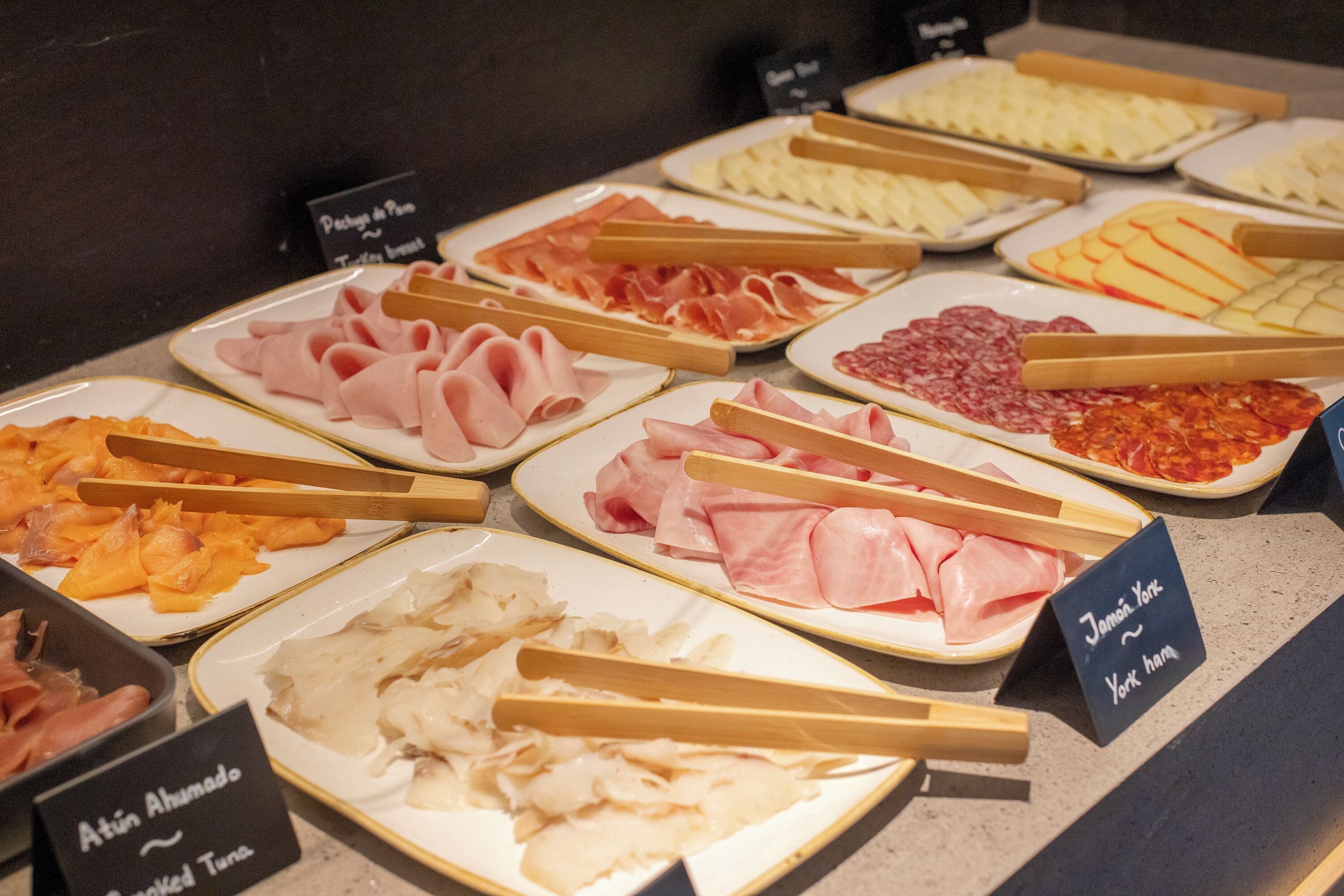 A variety of meats and cheeses displayed on plates with wooden tongs, including labels for 'Atún Ahumado' and 'Jamon Yori'.