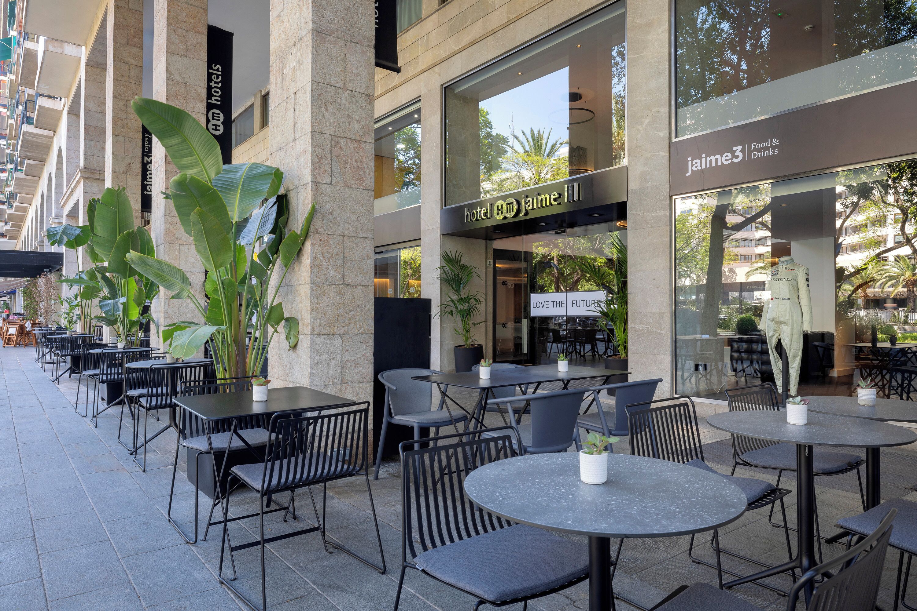 An outdoor cafe with tables and chairs, a large potted plant, and a building with a sign for 'jaime3 Food & Drinks'.