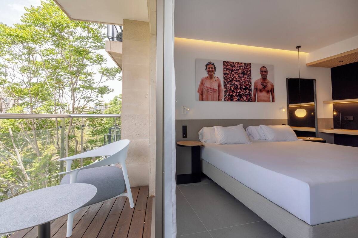 A modern hotel room with a balcony, a bed, a small table, and artwork on the wall.
