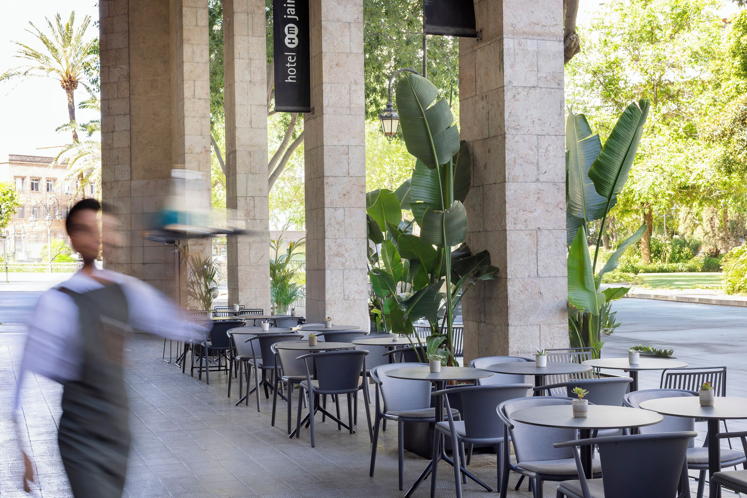 Un área de café al aire libre con mesas y sillas redondas, grandes macetas con plantas y una estructura de columna de piedra.