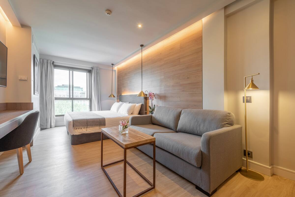 A modern hotel room with a wooden floor, a grey sofa, a wooden coffee table, a desk with a chair, and a large window with curtains.