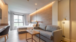 A modern hotel room with a wooden floor, a grey sofa, a wooden coffee table, a desk with a chair, and a large window with curtains.