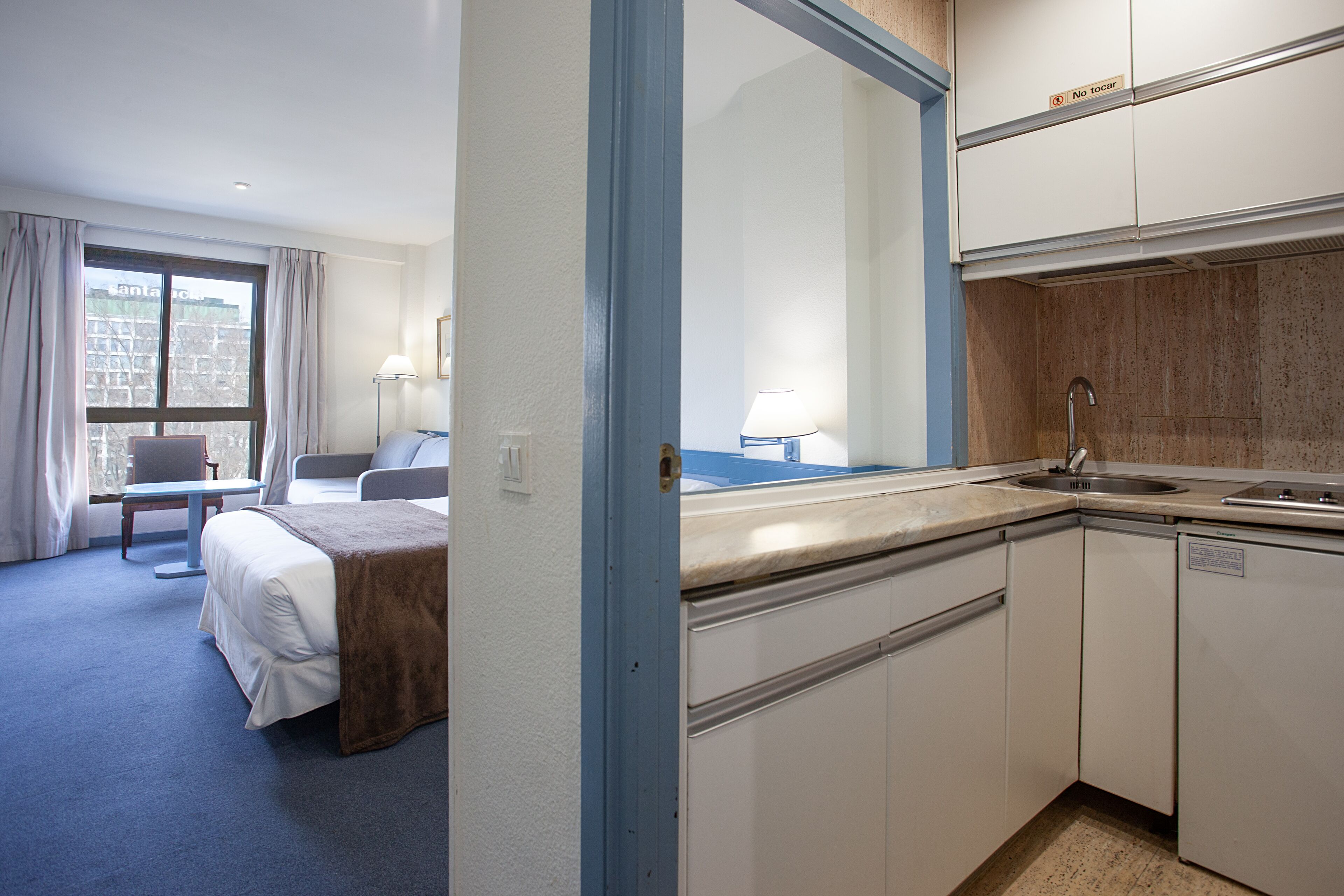 A hotel room with a bed, a desk, a chair, a window with curtains, a sink, and a mirror.