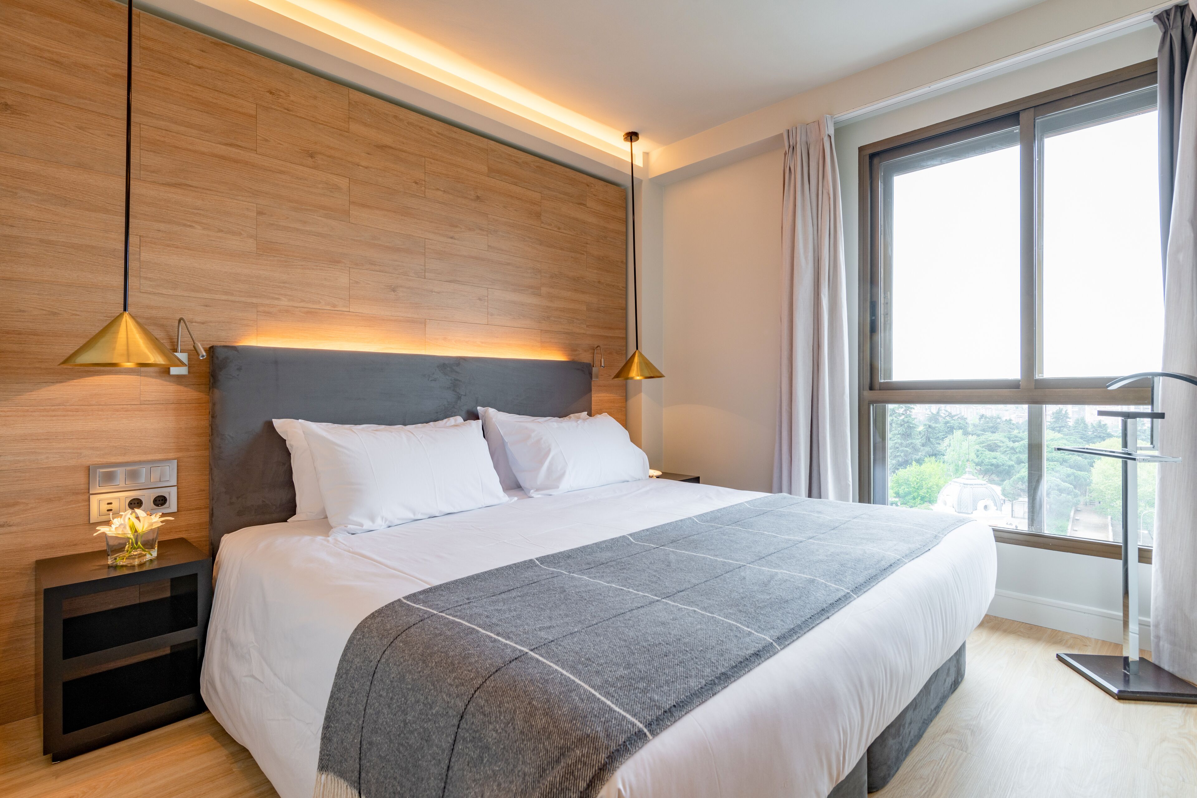 A modern bedroom with a large bed, wooden headboard, bedside lamps, and a view of greenery through the window.