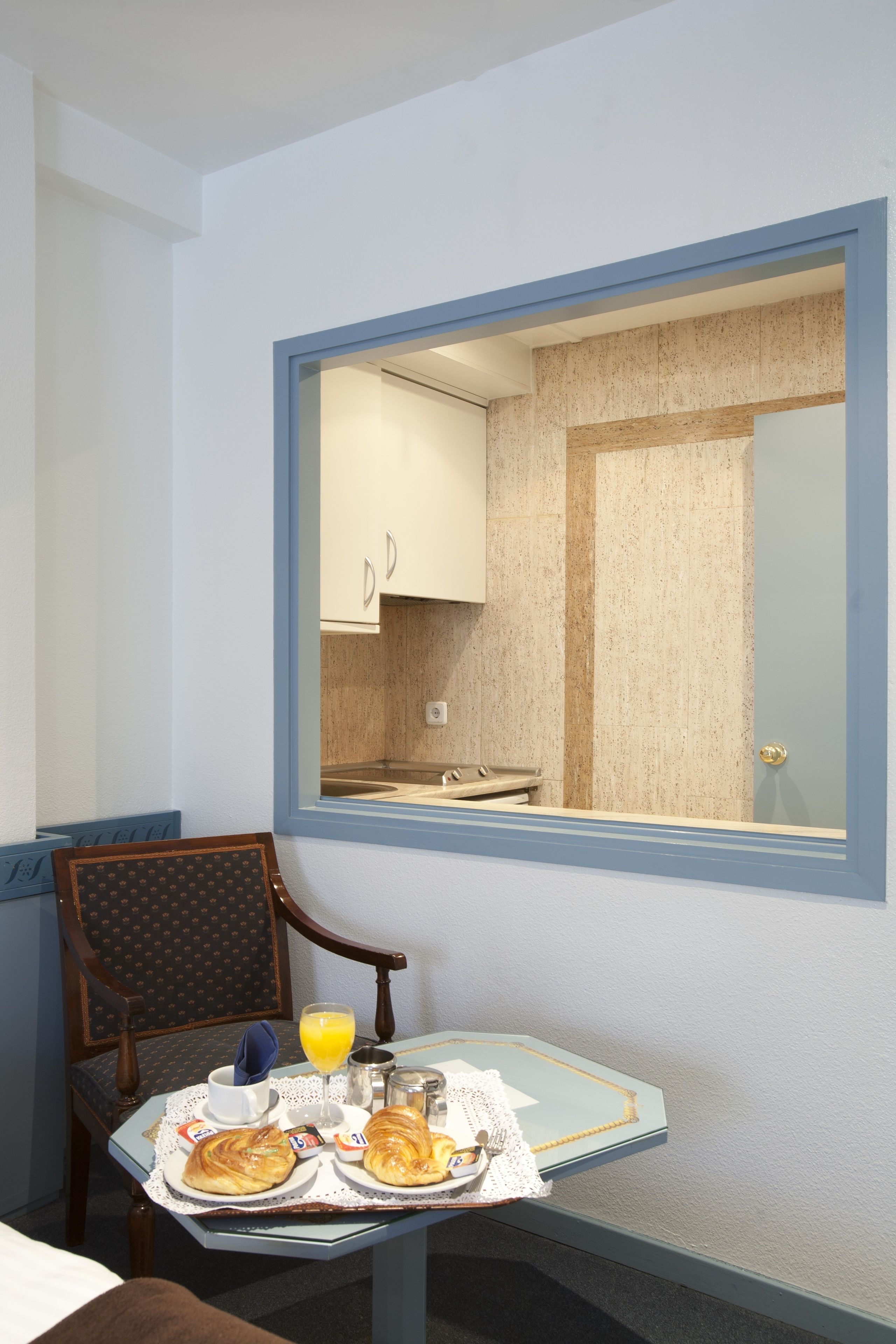 Una mesa de desayuno con cruasanes, un vaso de zumo de naranja y una taza, en una habitación con vistas a una cocina a través de una ventana.
