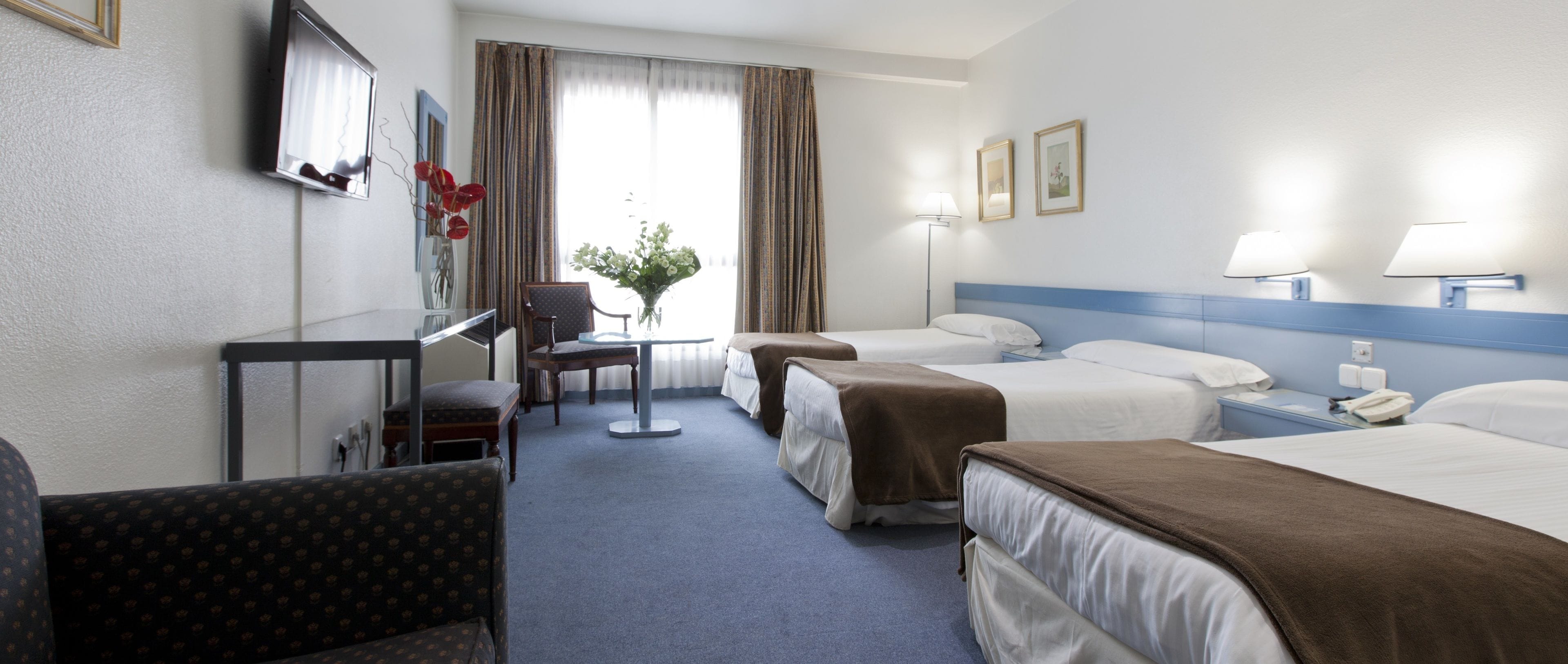 A hotel room with two beds, a desk, a chair, and a small table with a vase of flowers.
