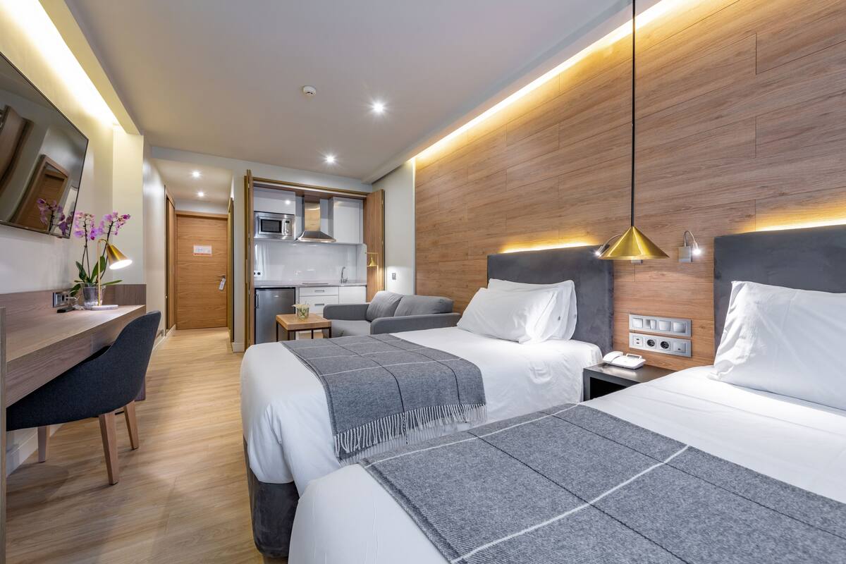 A modern hotel room with two beds, a desk, and a kitchenette.