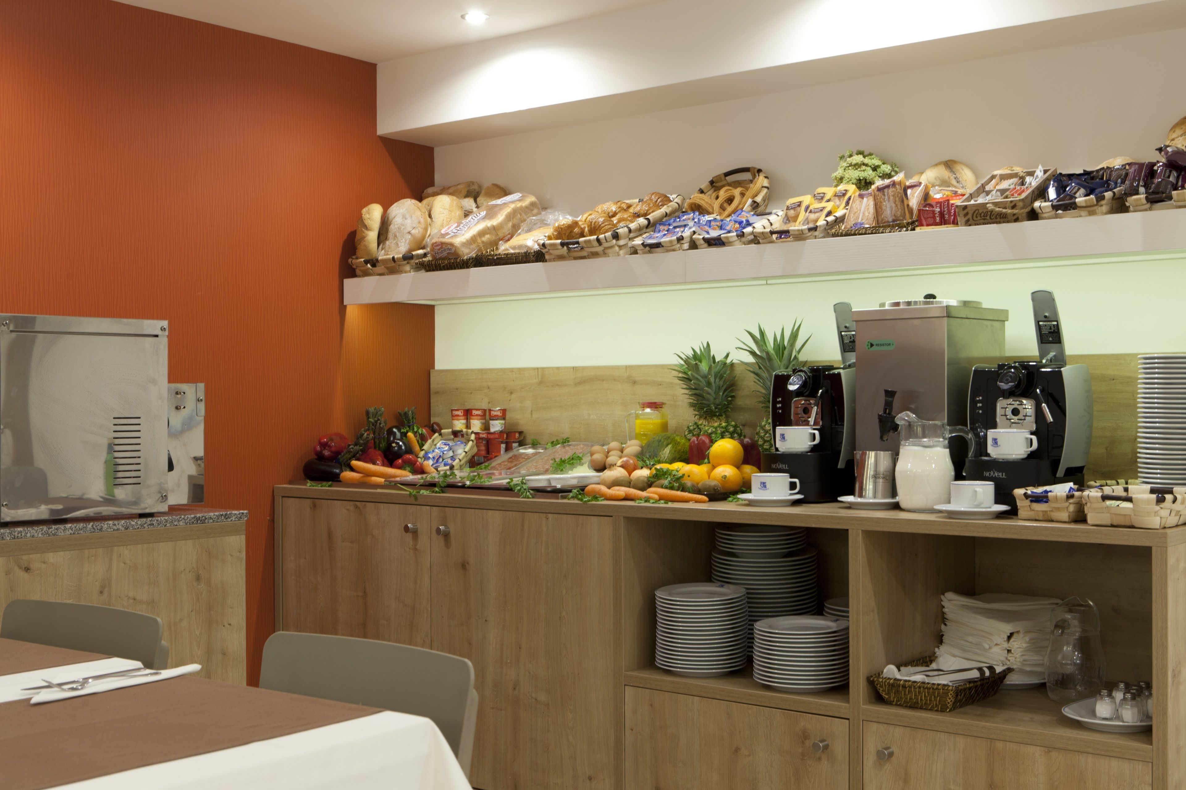 A breakfast bar with various breads, pastries, fruits, and beverages. There are stacks of plates and a coffee machine.