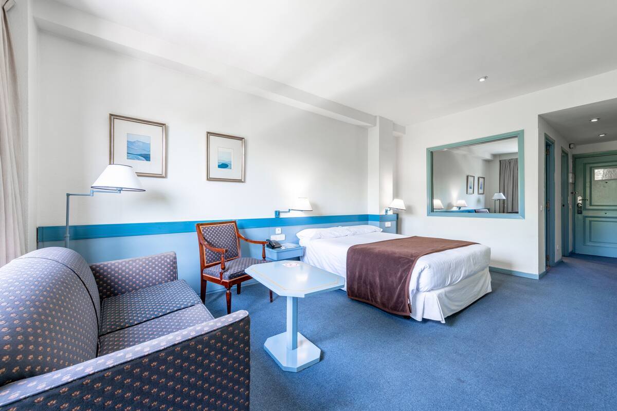 A hotel room with a bed, a chair, a small table, and two framed pictures on the wall.