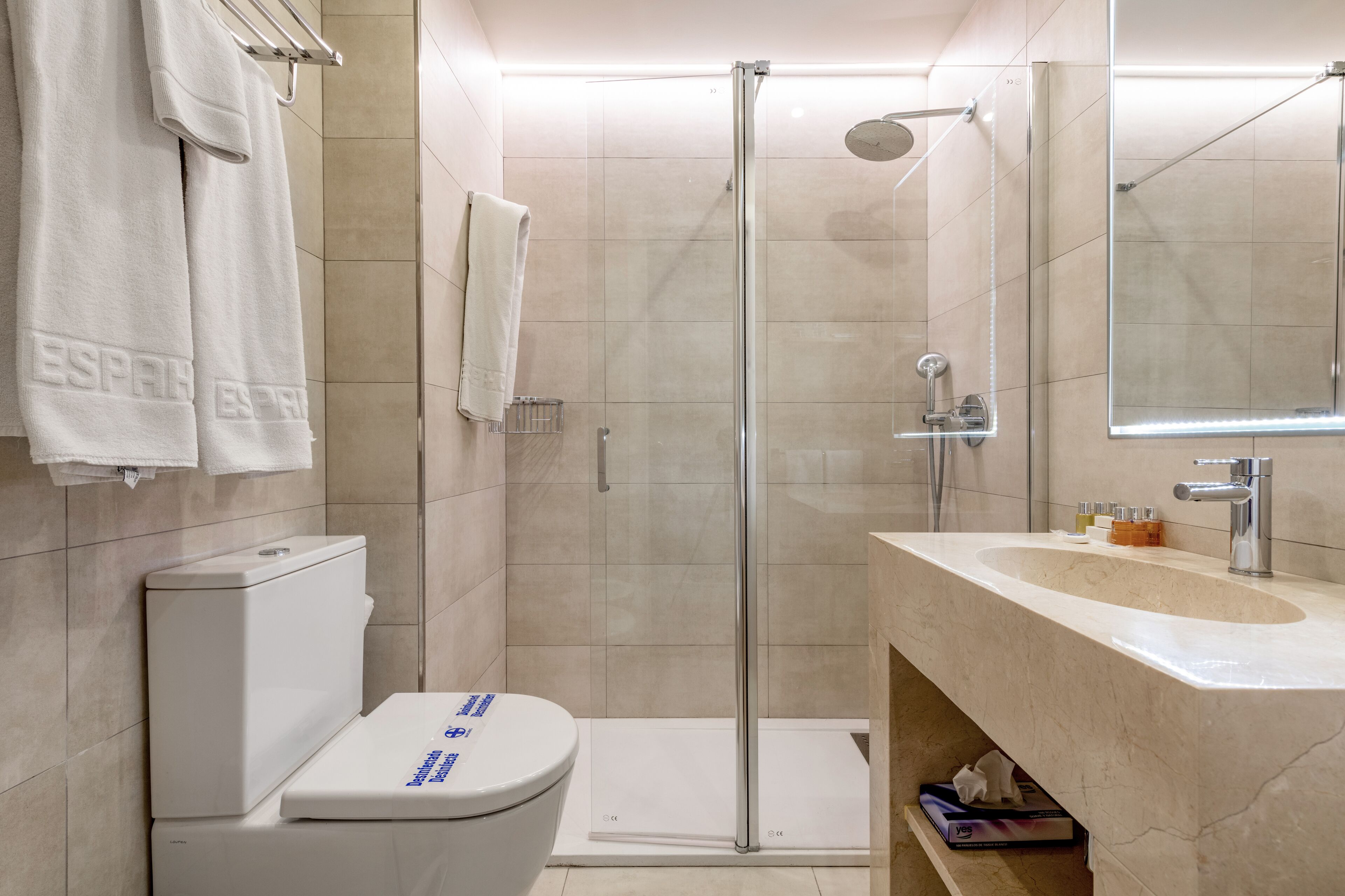 Un baño moderno con ducha envuelta en vidrio, inodoro blanco, lavamanos con espejo y toallas en un perchero.