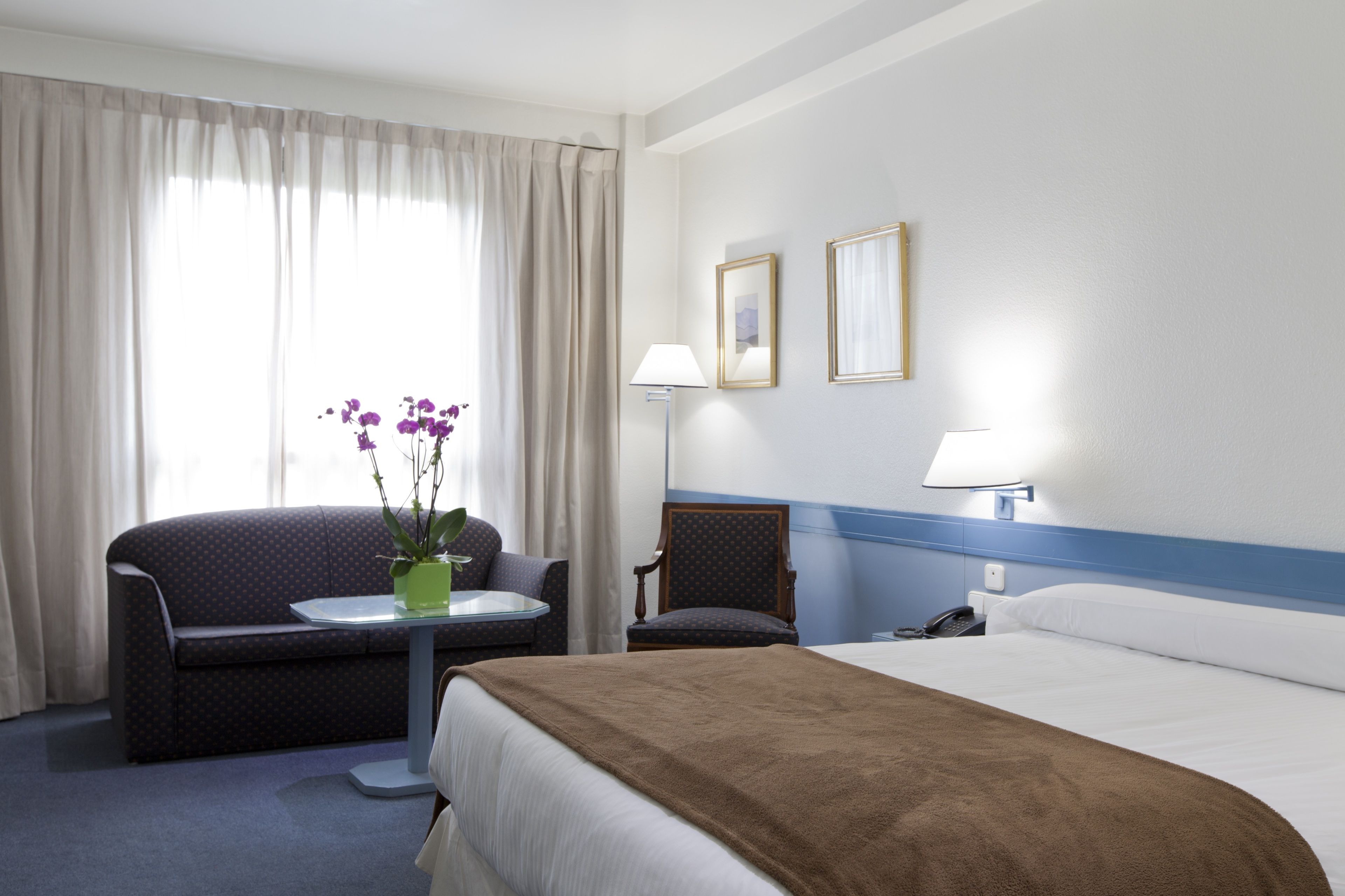 A hotel room with a bed, a sofa, a small table with a vase of flowers, a chair, and two wall-mounted lamps.