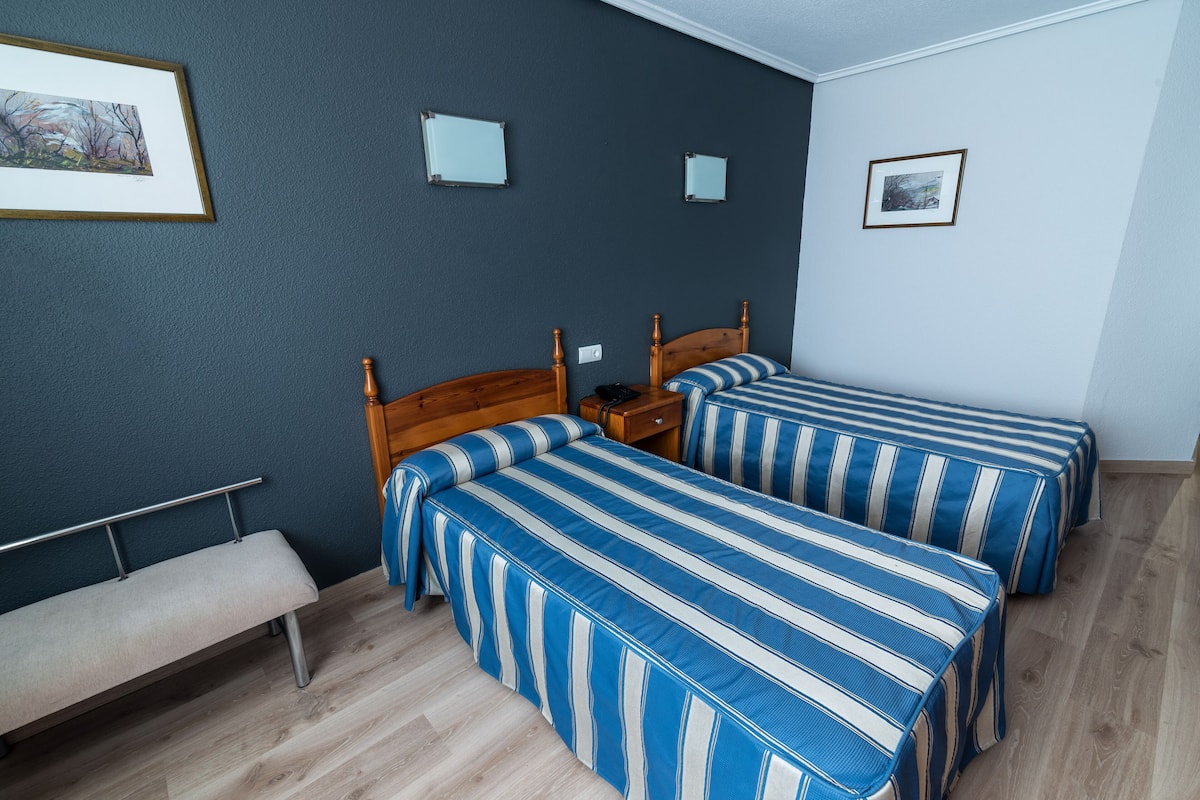 Habitación con dos camas, ropa de cama a rayas, un banco de madera y una pequeña mesa.