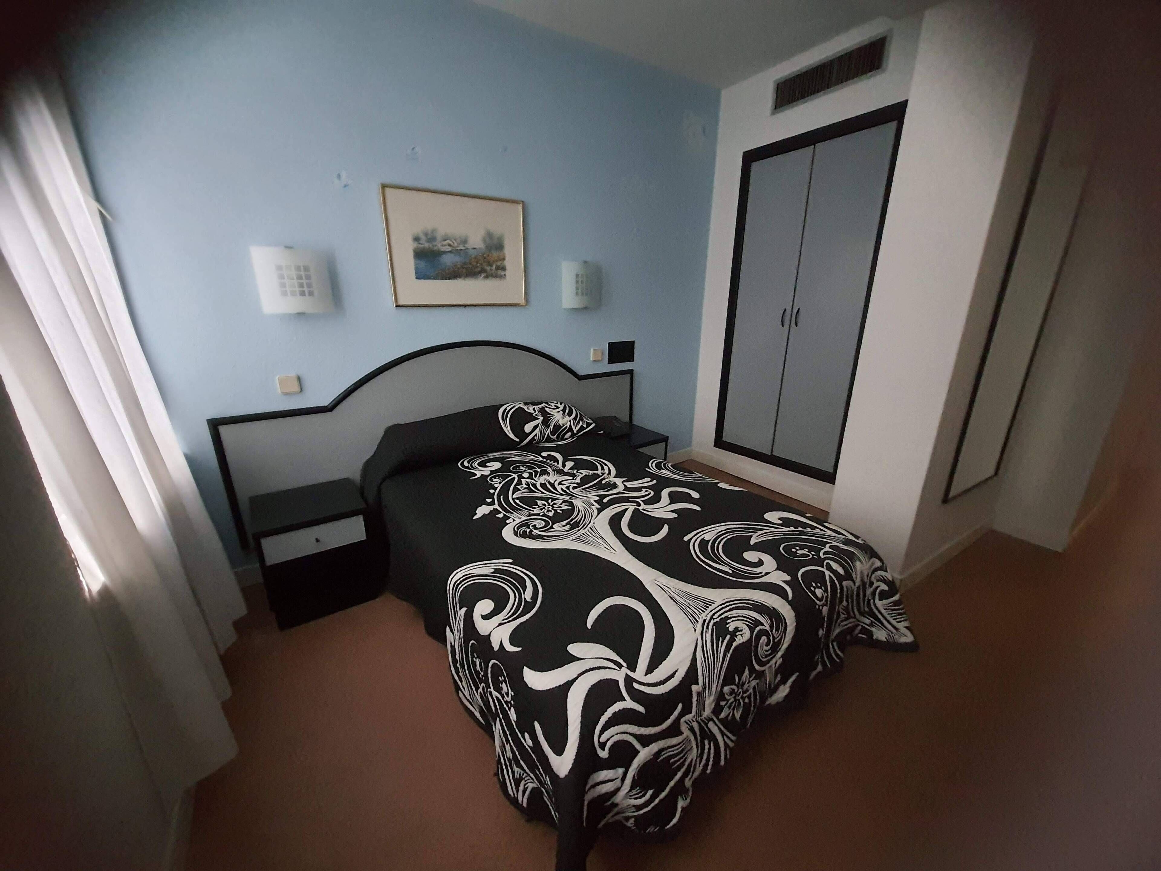 Un dormitorio con cama, armario, mesita de noche, un cuadro enmarcado en la pared y ventana con cortinas.