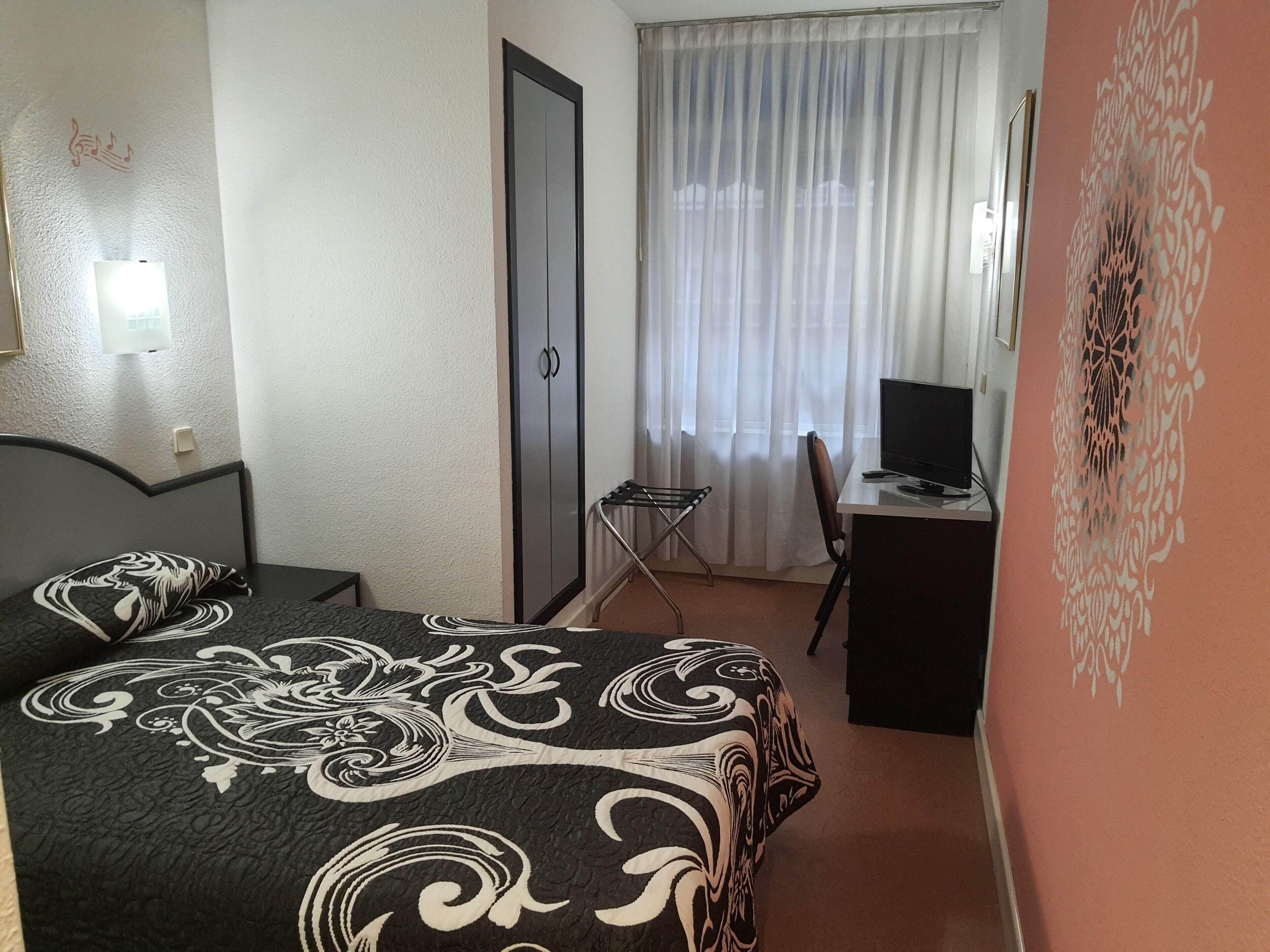 A bedroom with a bed, a desk with a computer, a chair, a wardrobe, and a window with curtains.