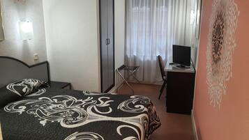 A bedroom with a bed, a desk with a computer, a chair, a wardrobe, and a window with curtains.