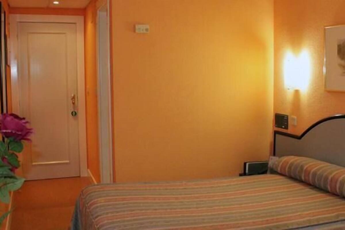 A hotel room with a bed, a nightstand, a wall-mounted lamp, and a door.