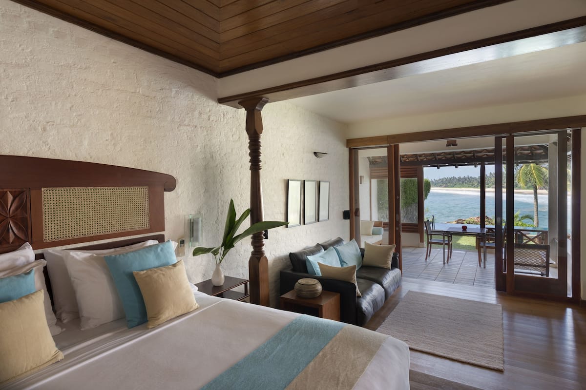 A bedroom with a large bed, a wooden headboard, a nightstand, a potted plant, and a view of the ocean through a glass door.
