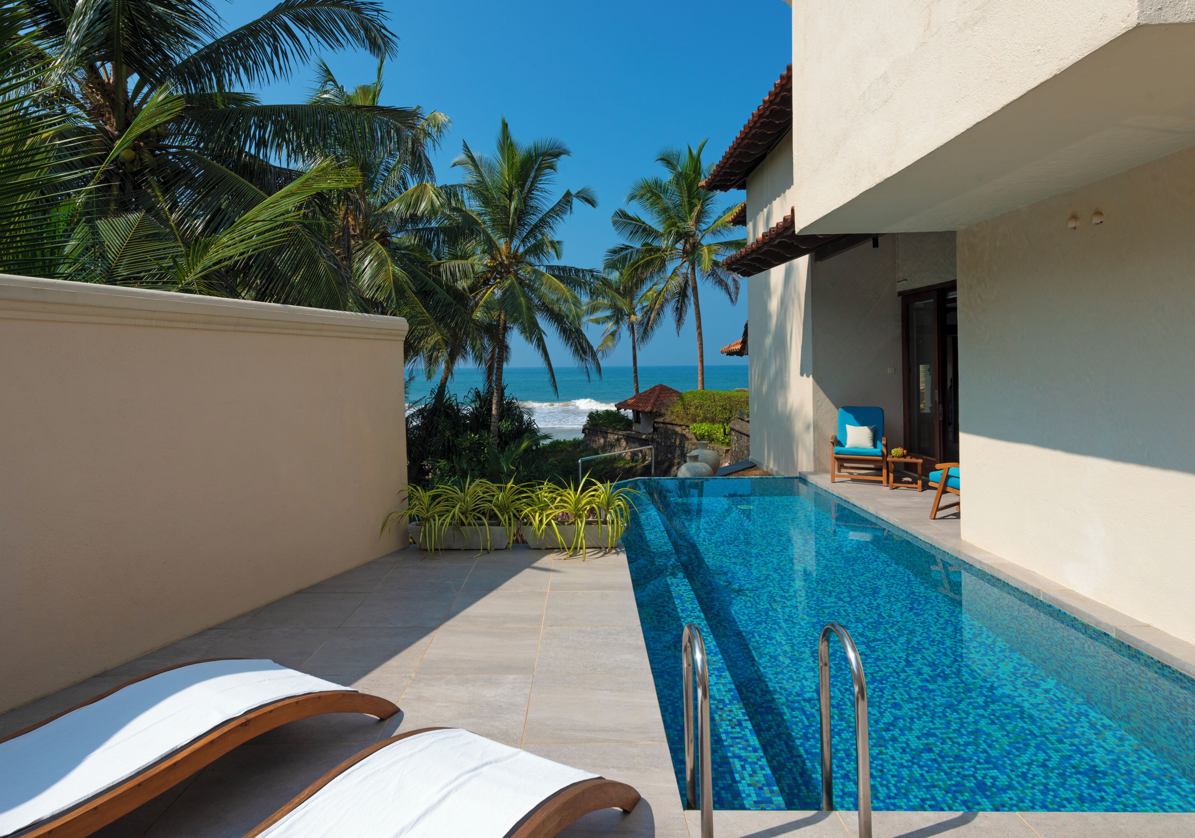 A swimming pool with lounge chairs, palm trees, and a view of the ocean.
