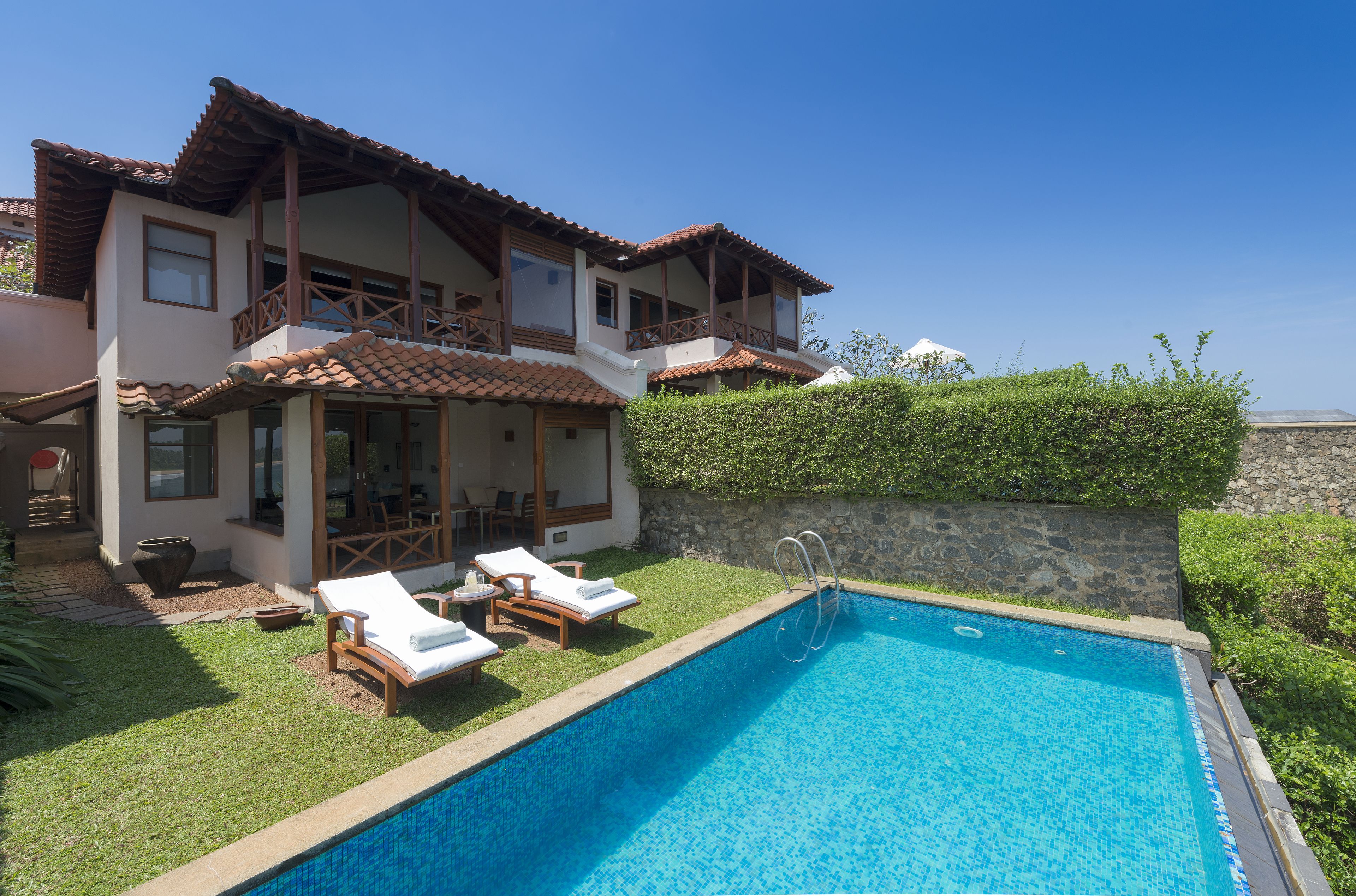 A two-story house with a swimming pool, outdoor furniture, and a stone wall.