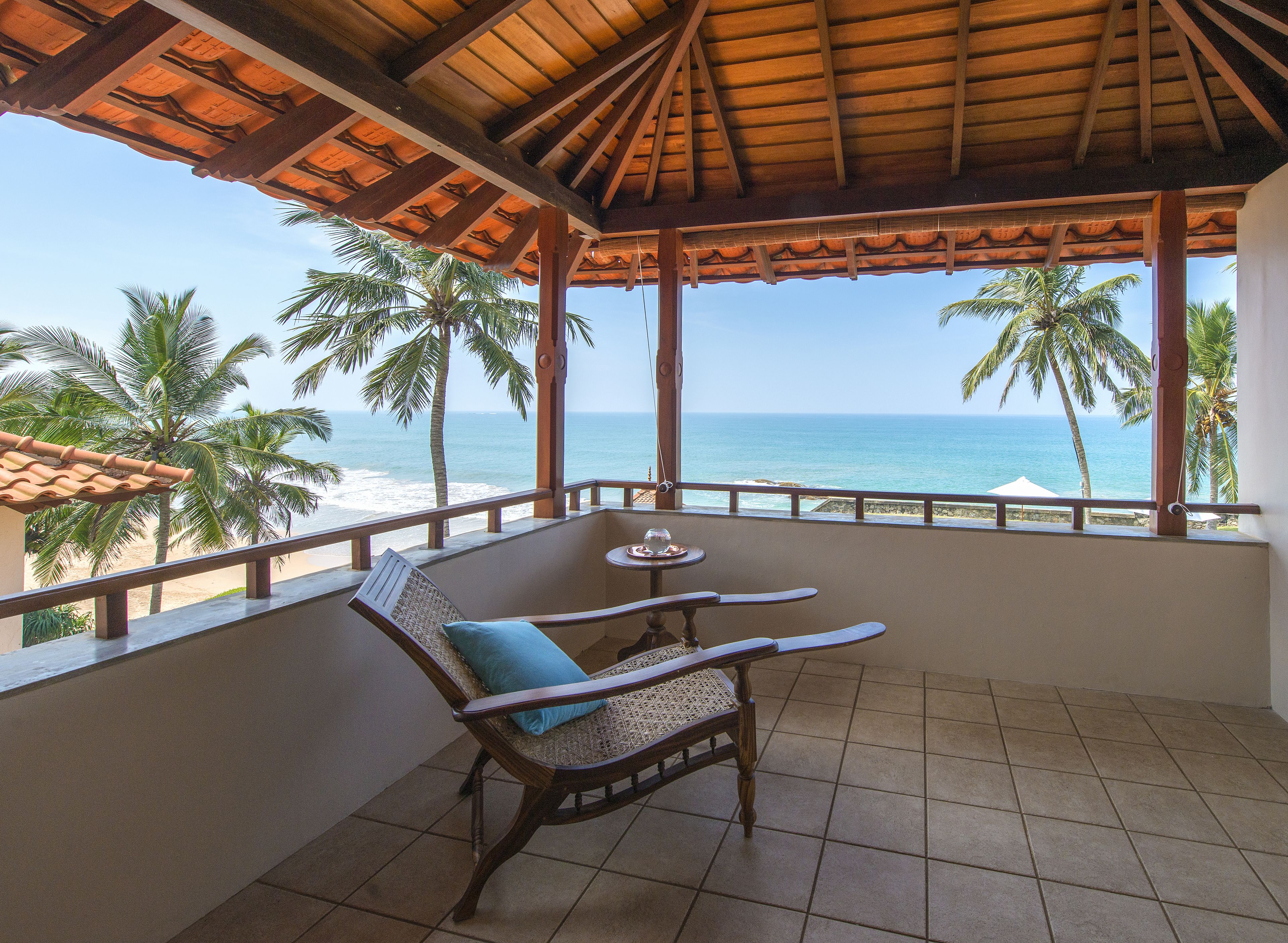 A balcony with a wooden table and two wicker chairs, overlooking a beach with palm trees and the ocean.