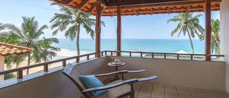 A balcony with a wooden table and two wicker chairs, overlooking a beach with palm trees and the ocean.