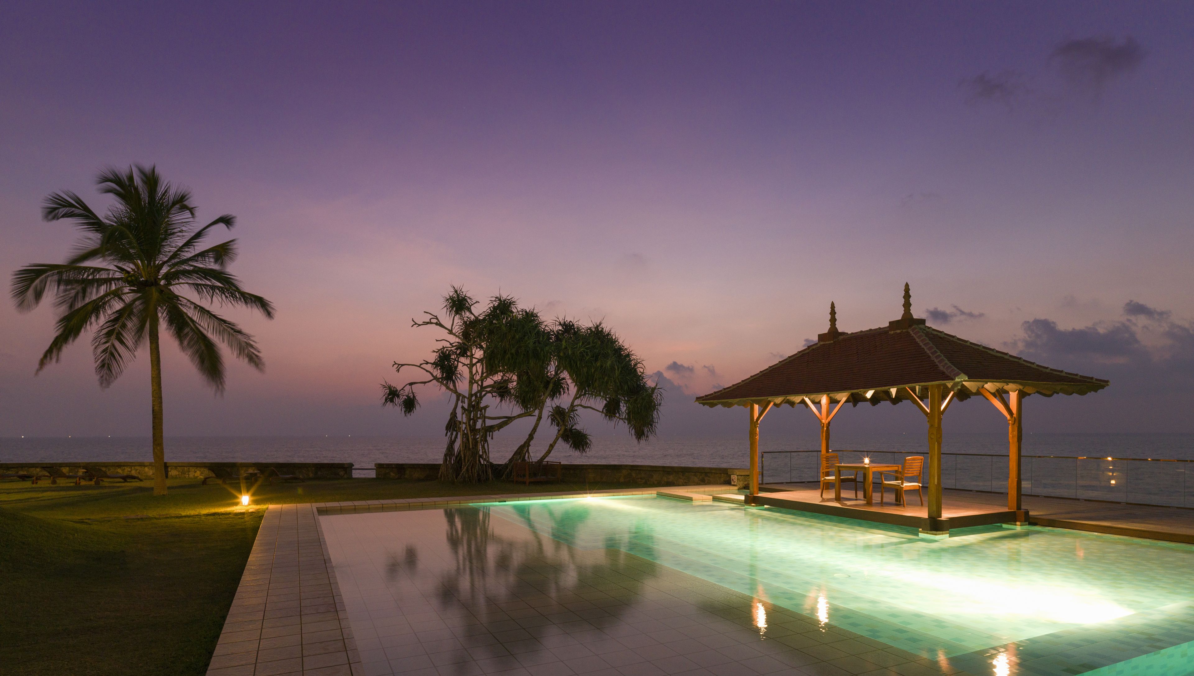 Ein Poolbereich mit einem Pavillon, Palmen und Blick auf das Meer im Abendlicht.