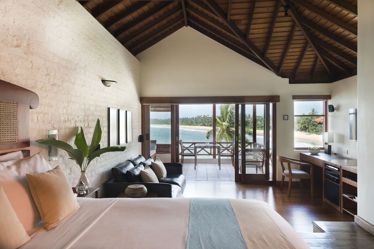 A spacious bedroom with a large bed, a sitting area with a sofa and chairs, a dining table, and a view of the ocean.