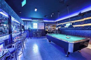 Una mesa de billar con taco y bolas, rodeada de taburetes de bar y una zona de bar con varias botellas.