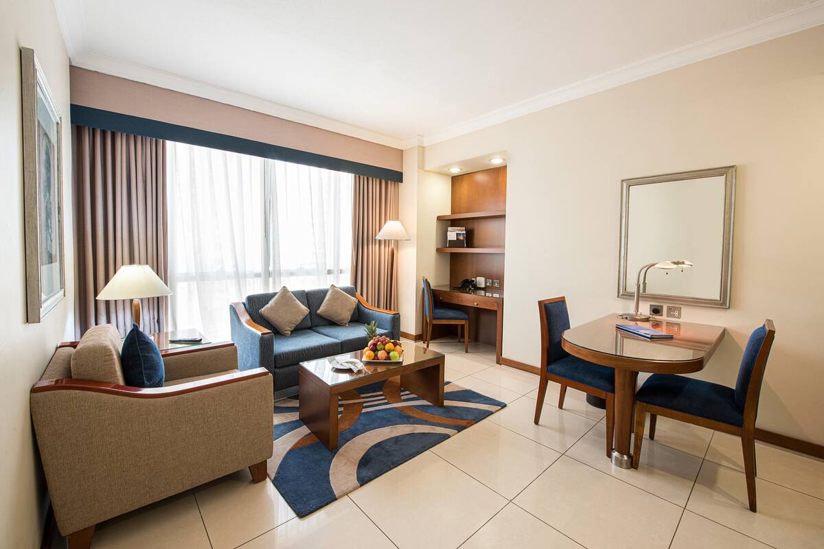 Ein Hotelzimmer mit einer Couch, einem Couchtisch mit Obst, einem Esstisch mit Stühlen, einem Schreibtisch und einem großen Spiegel.