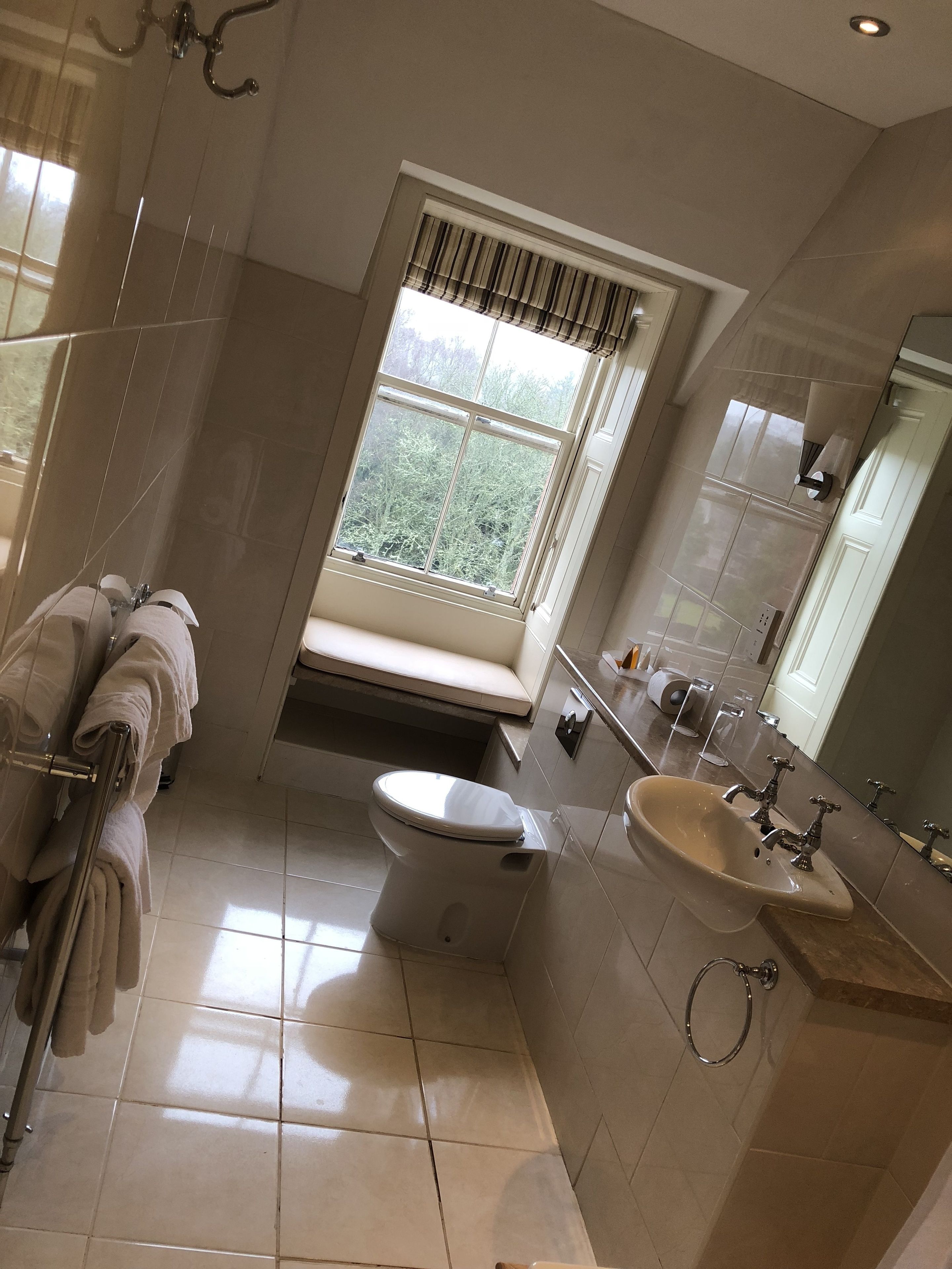 Une salle de bain comprenant des toilettes, un lavabo et une douche. On y trouve une fenêtre avec des stores, un porte-serviettes et un miroir.