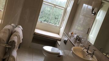 Une salle de bain comprenant des toilettes, un lavabo et une douche. On y trouve une fenêtre avec des stores, un porte-serviettes et un miroir.