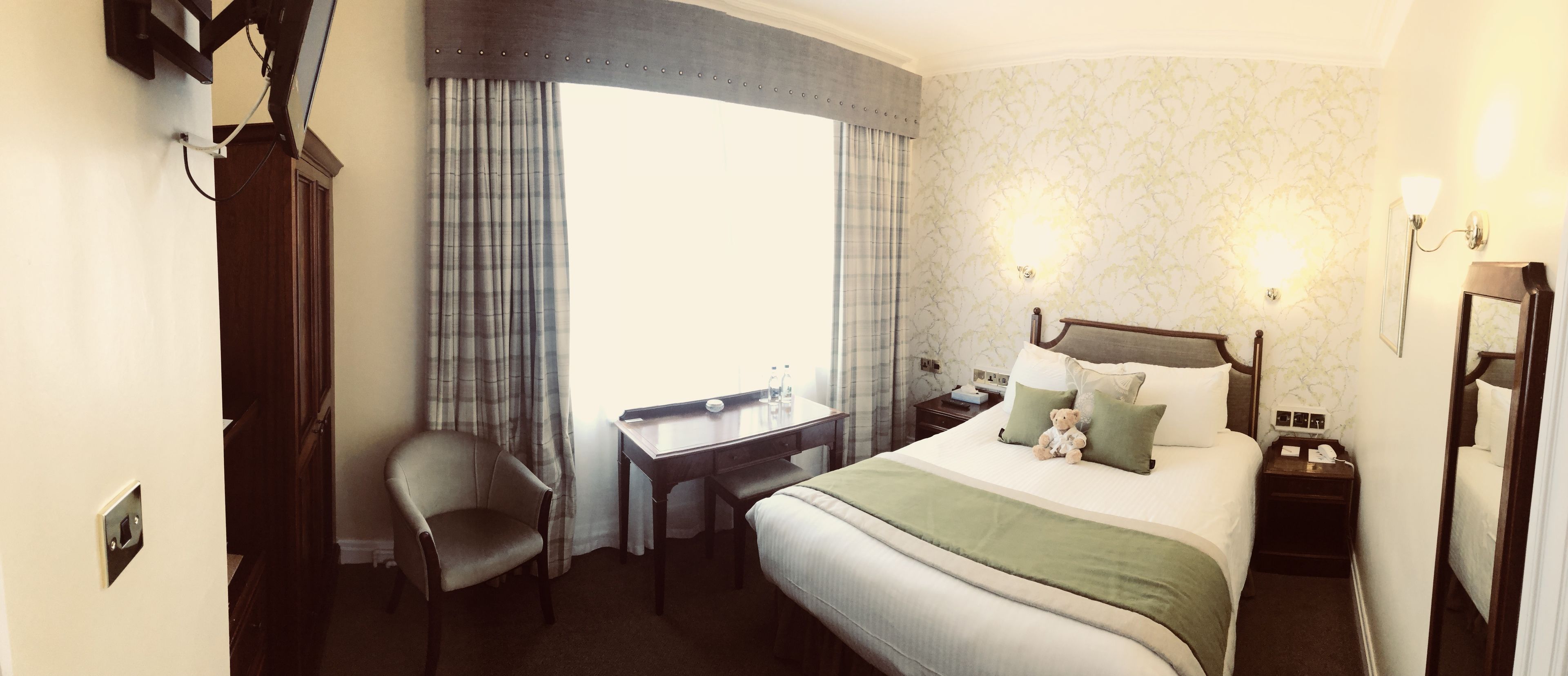 A hotel room with a bed, a desk, a chair, a nightstand, and a window with curtains.