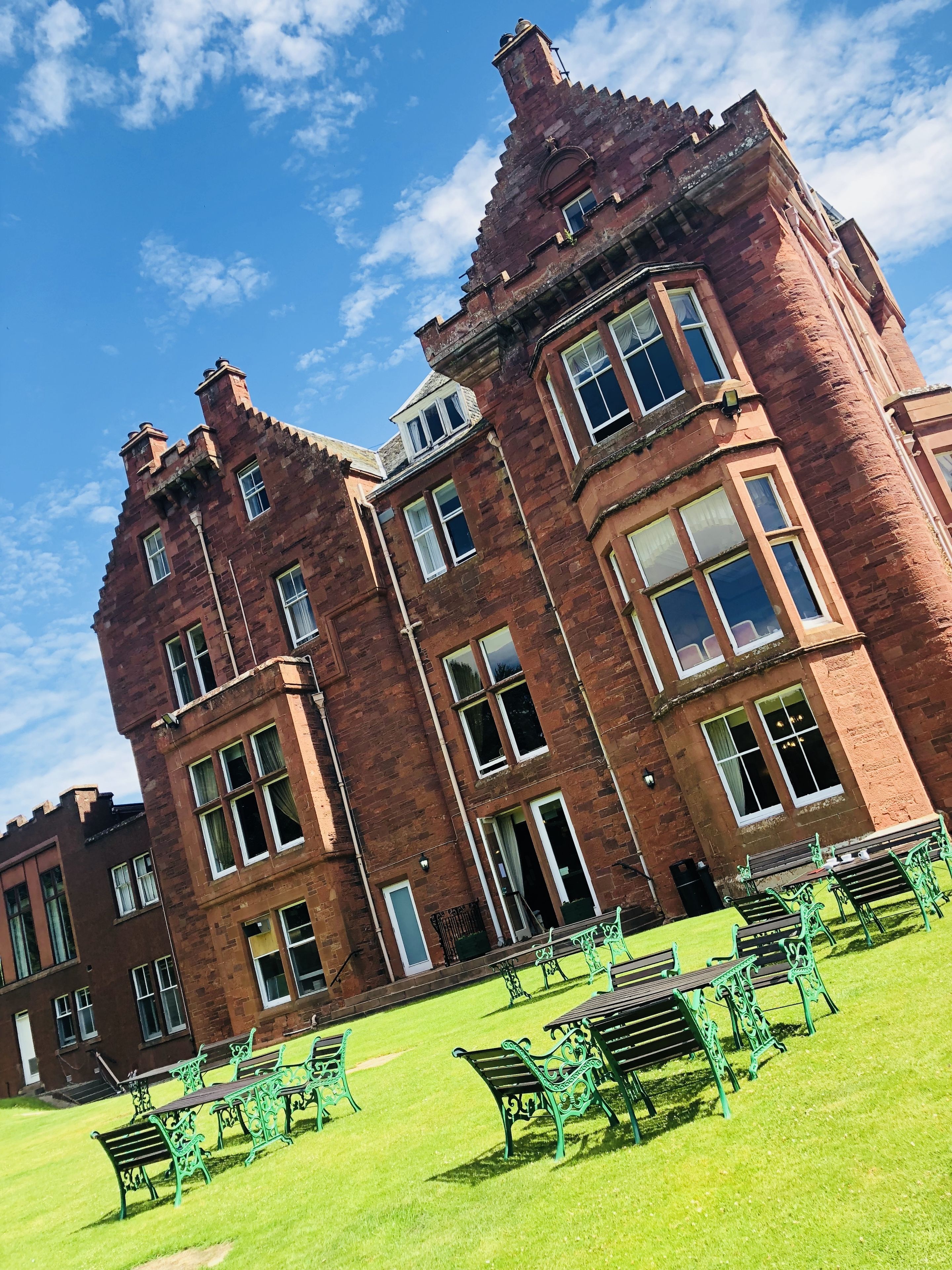 A large, historic red brick building with multiple windows and a lawn with green metal benches.