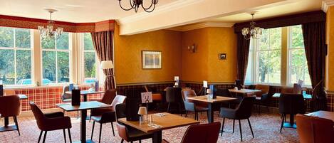 A hotel dining area with tables and chairs, large windows with curtains, and a chandelier.