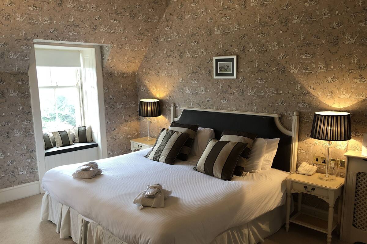 A neatly made bed with pillows and a blanket, a bedside table with a lamp, and a window with a view.