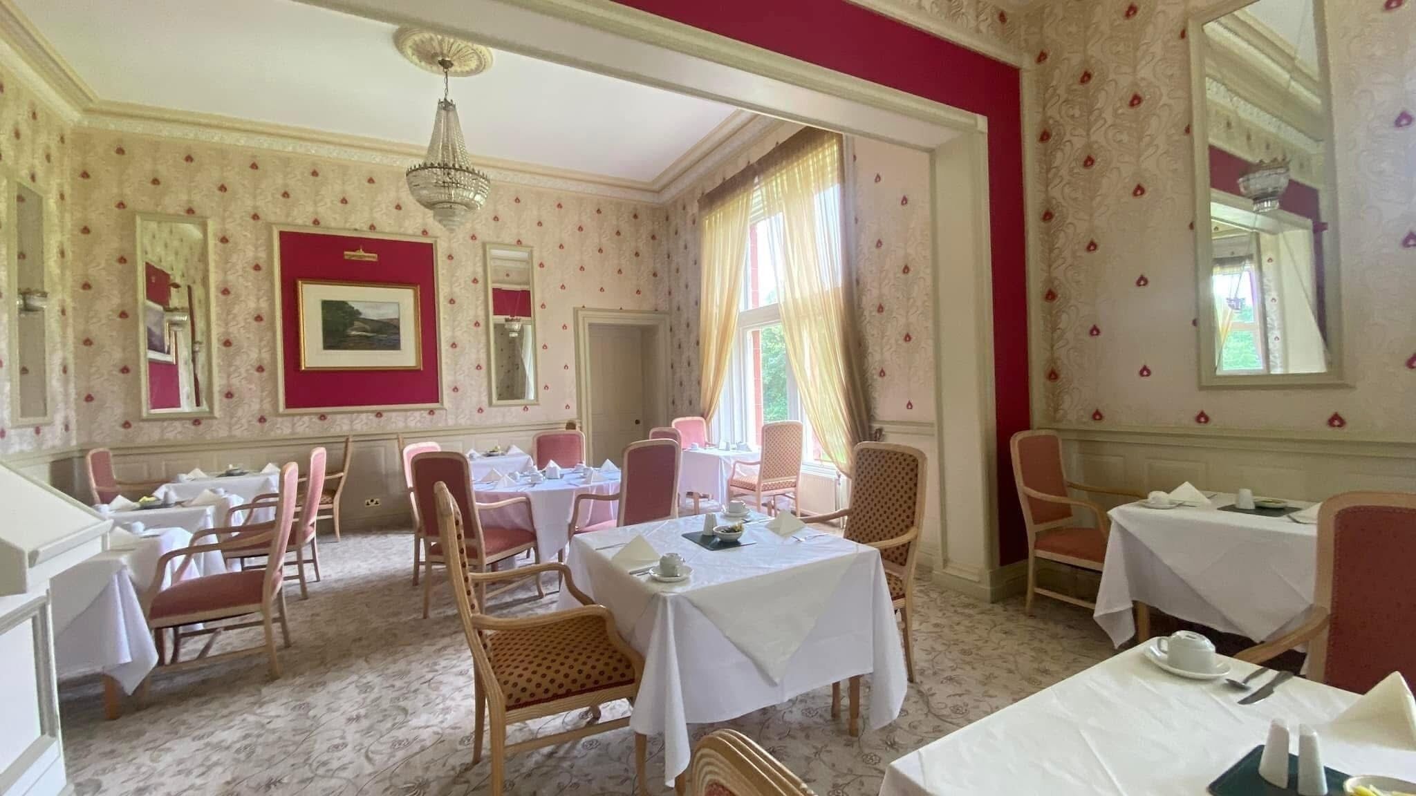 A dining room with multiple tables and chairs, a chandelier, and patterned wallpaper.