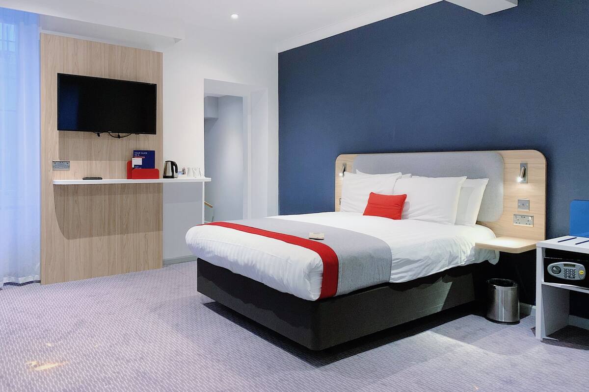 Una habitación de hotel moderna con una cama grande, un televisor de pantalla plana montado en la pared, un escritorio pequeño con una silla y un cesto de basura.